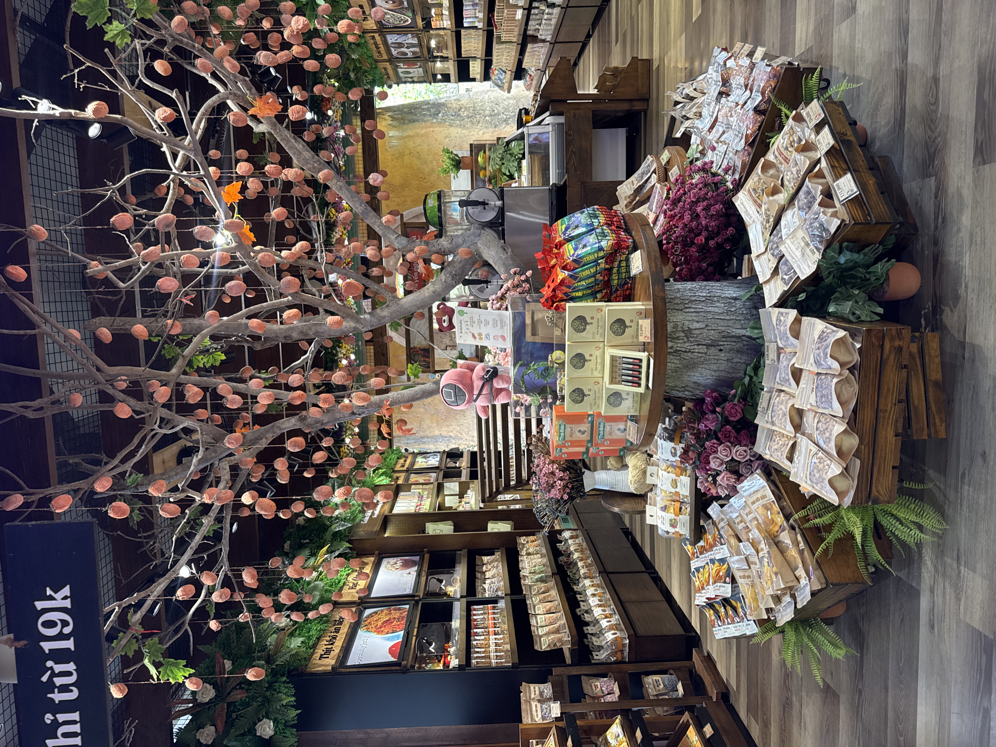 The image captures the interior of a commercial establishment, likely a store in Đà Nẵng, Vietnam, as indicated by the Vietnamese language signage. The space is well-lit and features a rustic-themed decor. Dominating the foreground on the left is a stylized tree-like structure adorned with numerous peach-colored spherical ornaments.
The store displays a variety of products on wooden shelves and tiered wooden stands. The shelves are lined with packaged goods, framed pictures of food, and various miscellaneous items. The wooden stands are laden with packaged snacks, nuts, and dried fruits. Floral arrangements and potted plants are interspersed among the merchandise, adding a touch of natural beauty.
A pink plush toy is visible on a shelf, and packaged goods with Vietnamese labels are prominently displayed. The floor is made of wooden planks, contributing to the overall rustic and inviting atmosphere of the store.