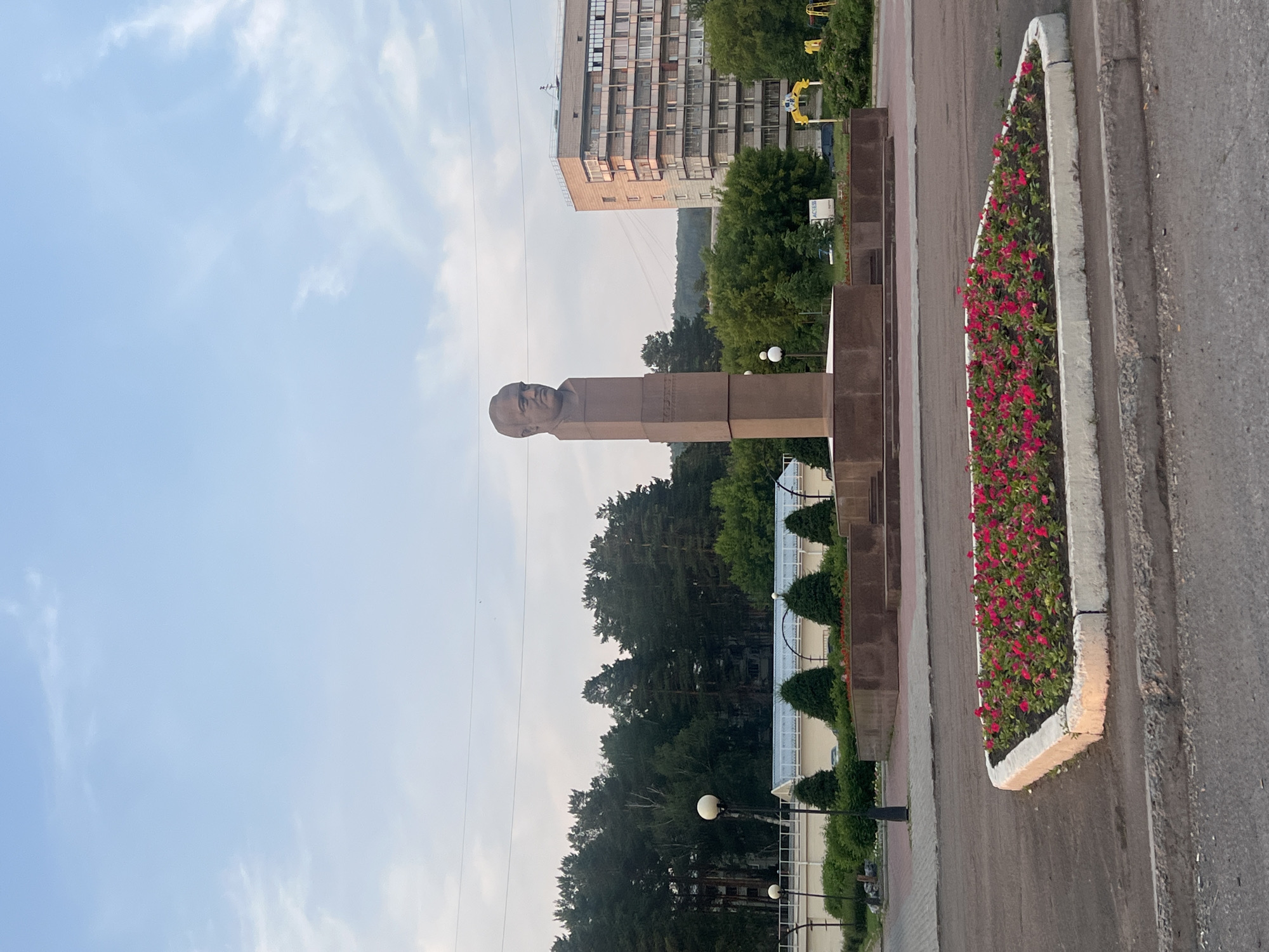 The image shows a bust of a man atop a tall, stone monument in the center of a paved public square. The monument is surrounded by a raised concrete flower bed filled with numerous small, red flowers and green foliage. A sidewalk borders the flower bed, and beyond that, a darker paved area, likely a road or parking lot. In the background, a multi-story apartment building with multiple balconies stands out against a backdrop of dense green trees. To the left of the monument, there are streetlights and a low building with a light-colored roof. The sky above is a pale blue with scattered white clouds. The location is identified as the memorial complex in honor of Academician Mikhail Lavrentyev in Zheleznogorsk, Russia. There are no people visible in the image.