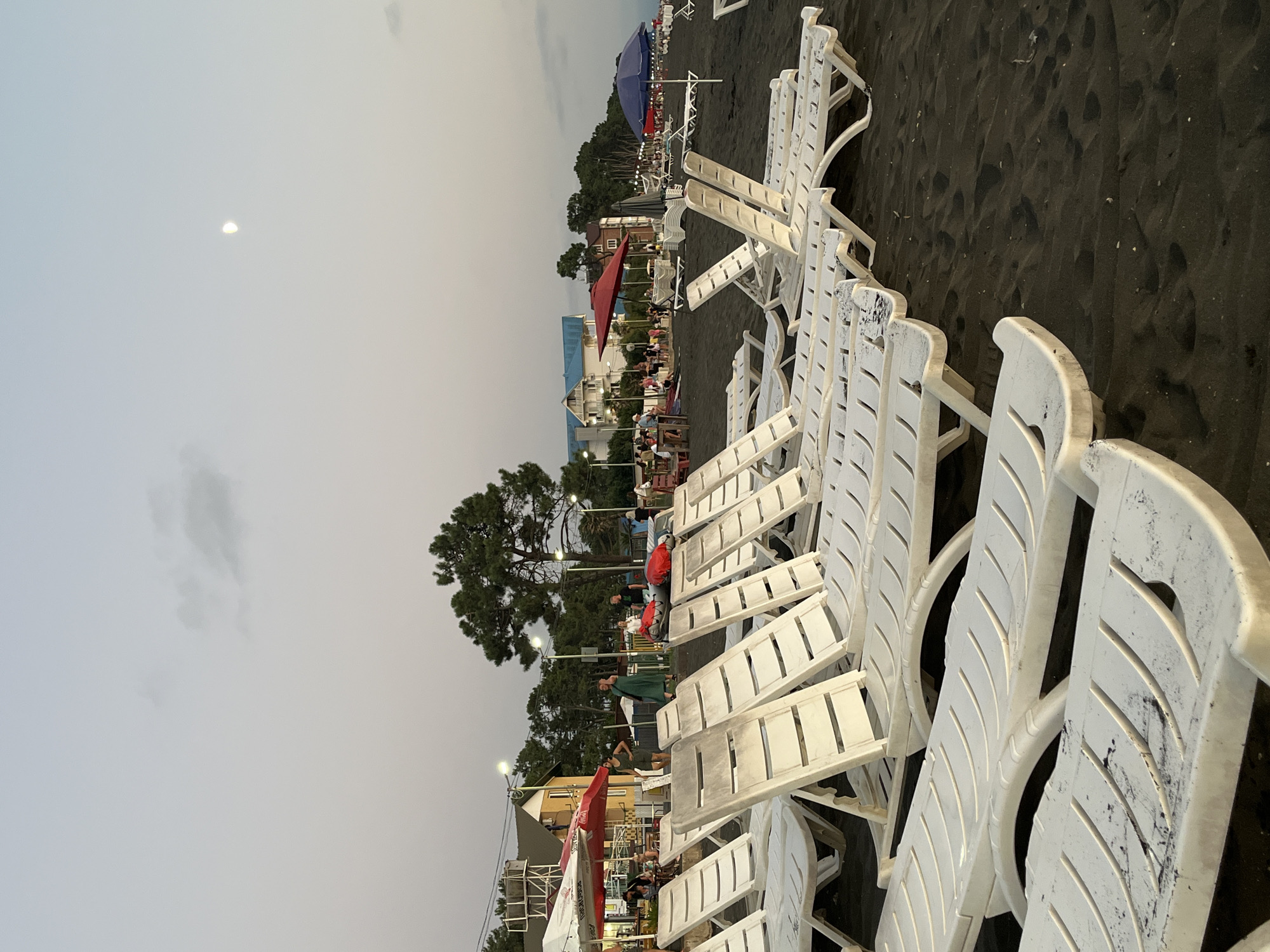 The image captures a view of Ureki, Georgia's famous magnetic sand beach at dusk. A line of white plastic lounge chairs dominates the foreground, arranged neatly on the dark, coarse sand. In the background, several people are visible, some standing and others seated at tables under umbrellas. Buildings, including houses and what appears to be a small cafe, are situated behind the beach, backed by a dense line of trees. The sky is a pale, hazy gray, with a sliver of the moon visible. The overall scene suggests a tranquil evening on the Black Sea coast.