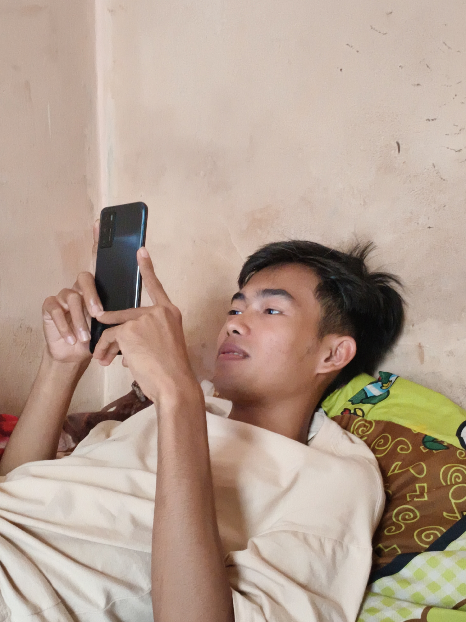 A young man with dark hair and a light complexion is lying on a bed, holding a smartphone in his hands. He is wearing a light-colored t-shirt. The wall behind him is textured and appears to be painted a light pinkish-beige color. To his right, a pillow with a green and brown pattern is visible. The overall scene suggests an indoor setting, likely a bedroom. The image does not contain enough information to determine a specific location within Cianjur, Indonesia.