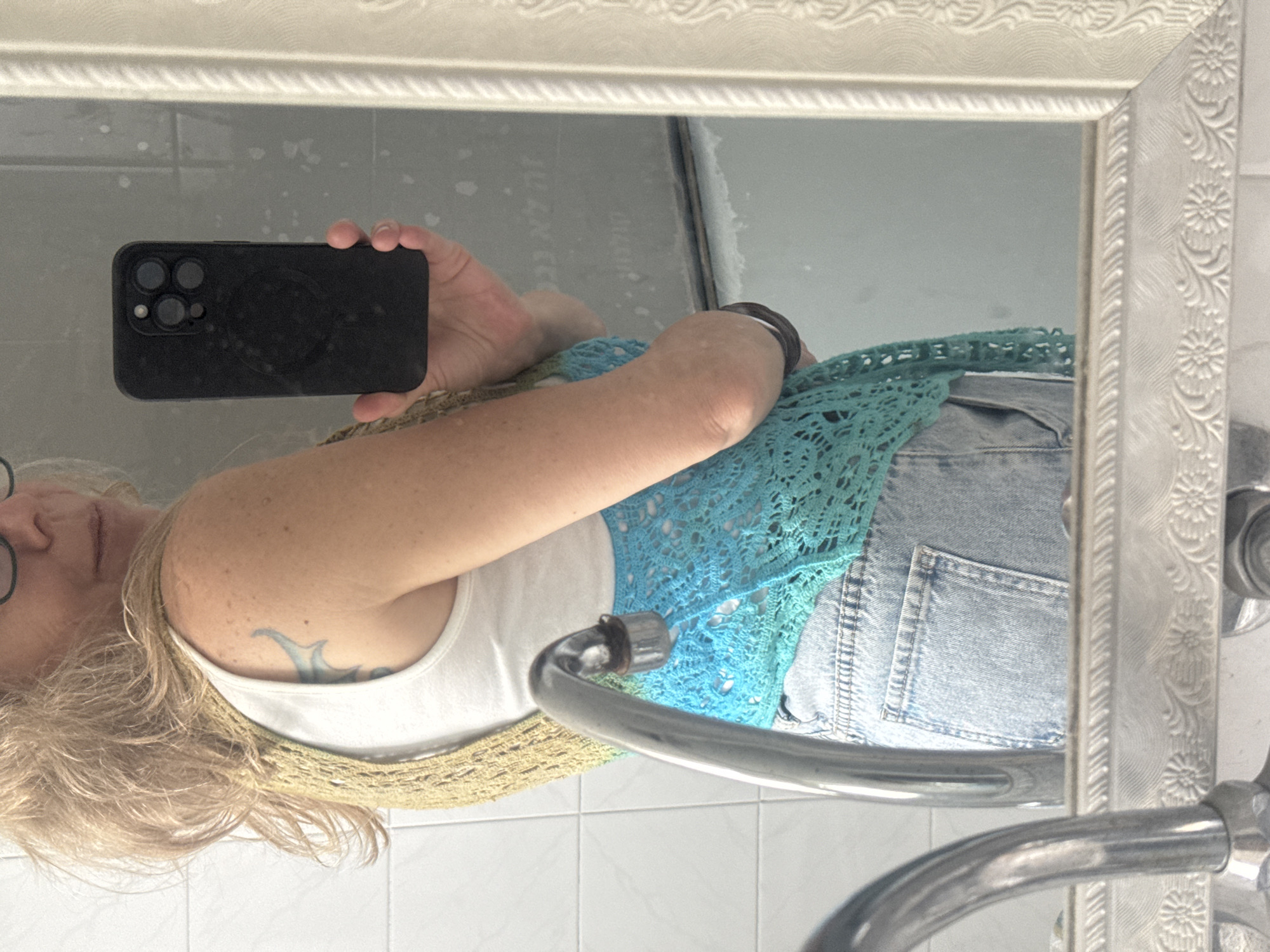 A selfie is taken in a bathroom in Tel Aviv, Israel. A blonde woman with glasses holds a smartphone in her right hand, facing a mirror with a white, floral-patterned frame. The mirror reflects the woman wearing a white tank top, a crocheted vest with gold and teal colors, and light blue denim shorts. The bathroom has white tiles. A silver faucet is positioned in front of the mirror.