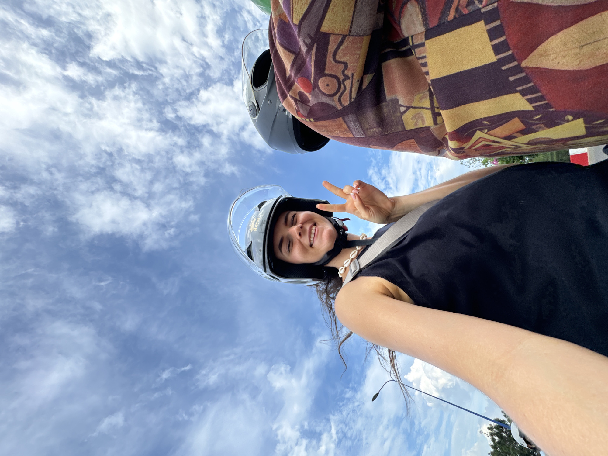 This low-angle photograph, likely a selfie taken during a scooter ride in Mũi Né, Vietnam, features two individuals against a vibrant blue sky filled with scattered white clouds. A smiling woman, positioned prominently, wears a black sleeveless top, a decorative shell necklace, and a dark grey helmet with a clear visor. She looks cheerfully at the camera while making a peace sign with her fingers. The word "Royal" is faintly visible on her helmet.
Partially visible above and behind her is a second person, presumably the driver, also wearing a dark helmet and a distinctive, earthy-toned jacket with abstract geometric patterns in shades of red, brown, and yellow. The upward perspective emphasizes the expansive sky, suggesting bright, clear daylight and good weather. Hints of green foliage and a roadside structure are visible towards the bottom right, grounding the joyful and casual scene. The overall mood is carefree and happy.