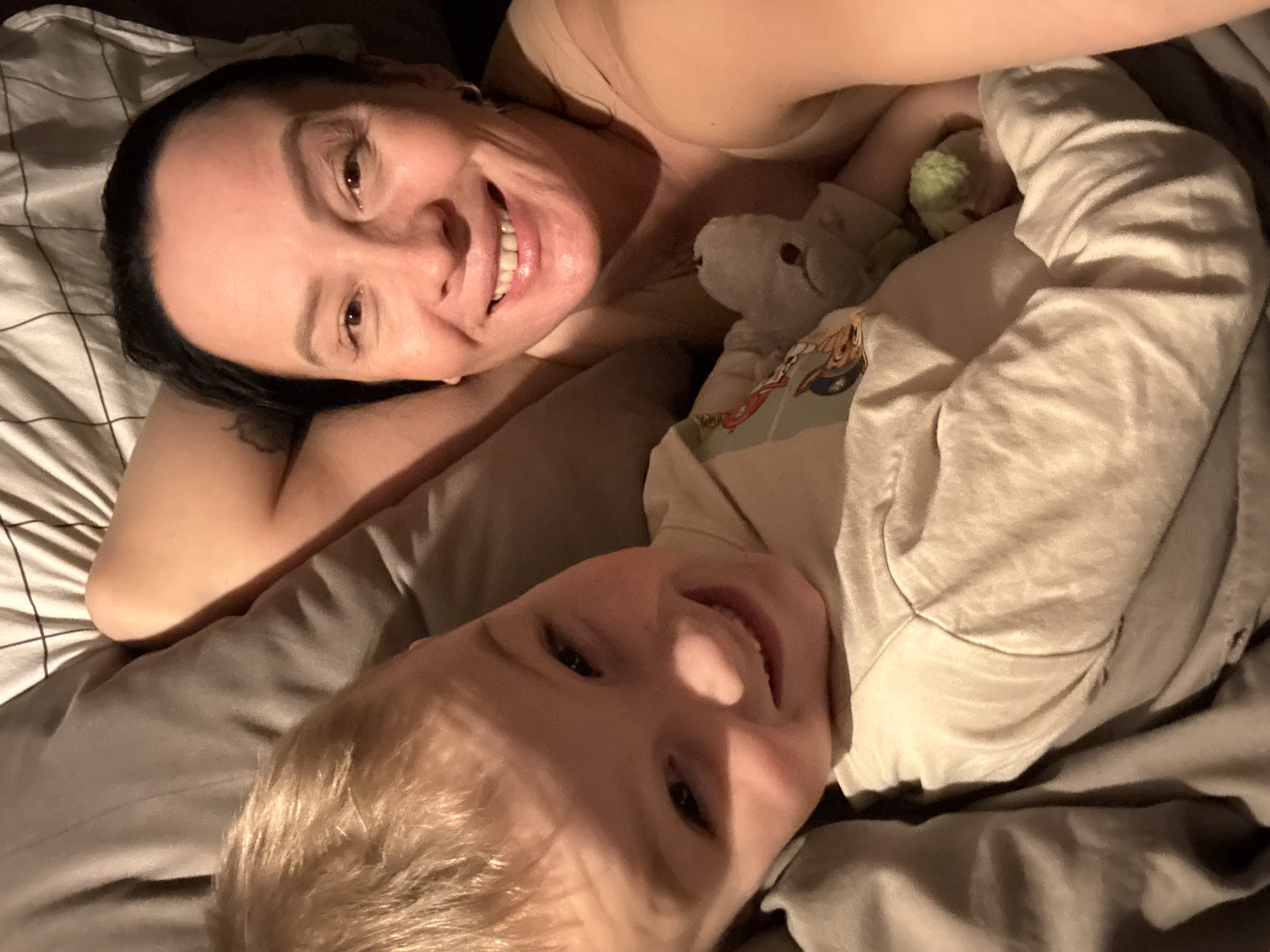 A selfie-style photograph shows an adult and a child lying on a bed with gray and white striped sheets. The adult, on the left, has dark hair and is smiling at the camera. The child, on the right, has blond hair and is also smiling. Between them, there is a white stuffed animal with a cartoon character shirt and another green stuffie. The lighting suggests that it is an indoor scene. The location is unidentifiable.