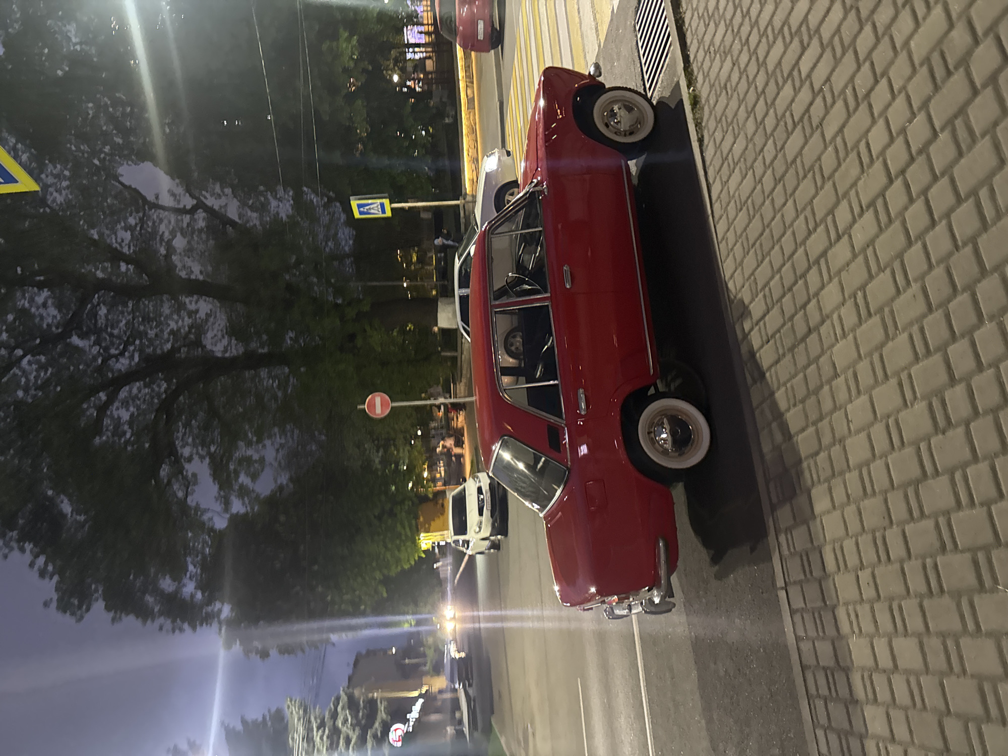 A red vintage car is parked on the side of a street next to a cobblestone sidewalk. The location is indicated by a pedestrian crossing sign visible nearby. The background features trees and buildings, with streetlights illuminating the evening scene. A white vehicle is parked in front of the red one. The sign "Komgaron" is visible in the distance suggesting the location within the city of Komgaron, Russia.