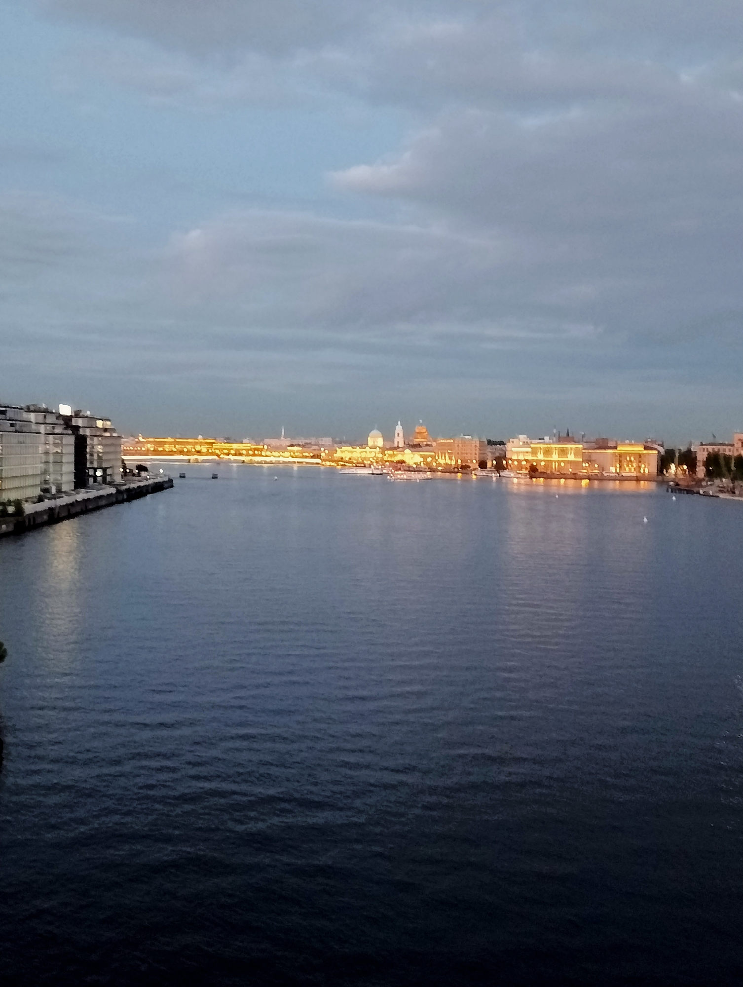 The image depicts a panoramic view of the Neva River in Saint Petersburg, Russia, at dusk. The left bank of the river features a modern, multi-story building with a glass facade. Across the river, the right bank is lined with historic buildings illuminated by warm yellow lights, suggesting a city center location, possibly the Palace Embankment area given the architectural style, which includes domes and classical facades. The sky is overcast with muted gray clouds, transitioning from a lighter blue near the horizon. The water of the Neva River is dark blue and shows ripples, indicating a slight current. Several small boats and distant lights are visible on the river and along the embankment. The overall atmosphere is calm and serene, characteristic of a twilight scene in a major European city.