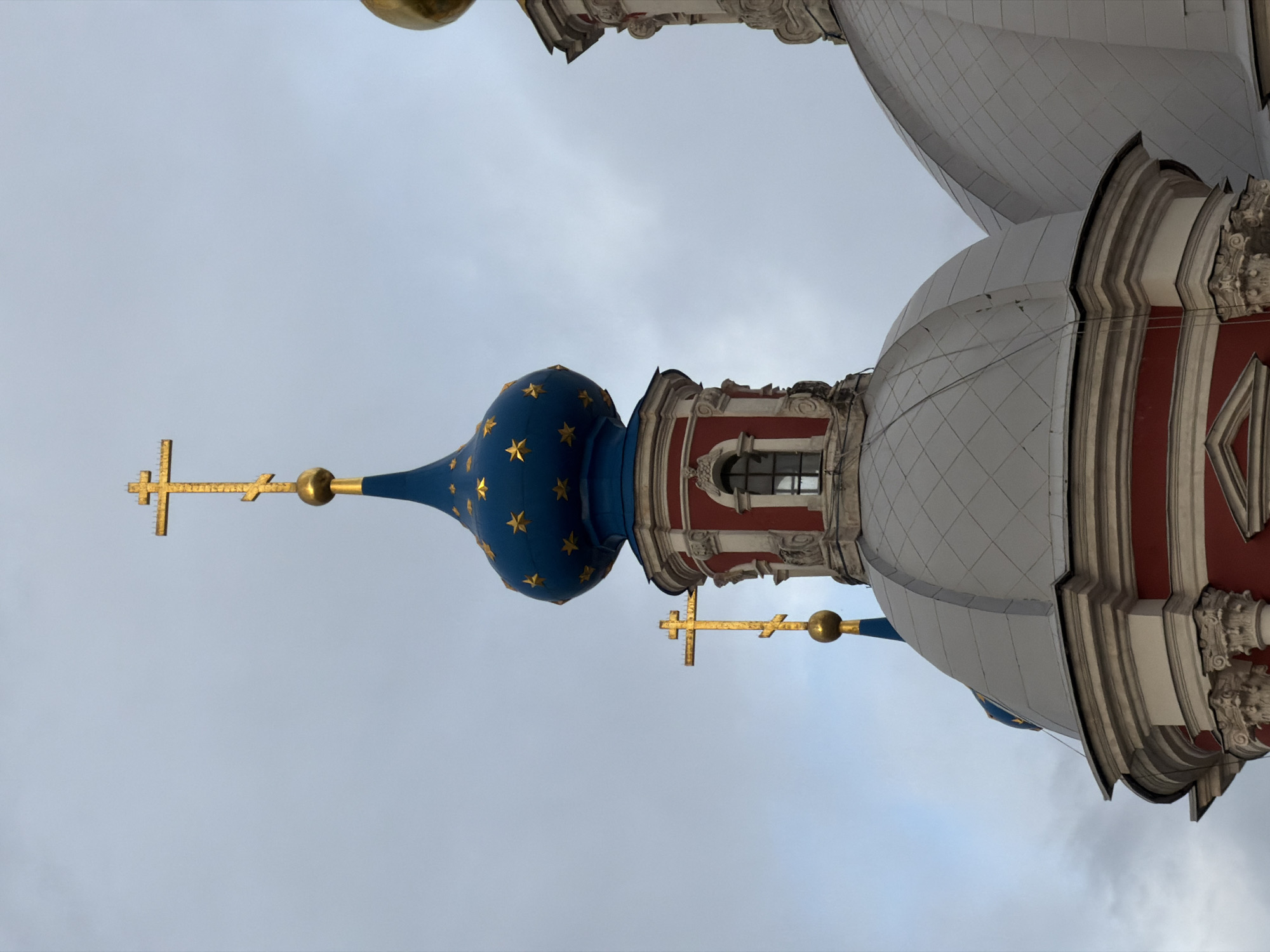 The image shows a view of the Novodevichy Convent in Moscow, Russia. The camera angle is looking upward towards the domes and crosses of one or more of the buildings. A large dome tiled in white and grey, trimmed in red and white, is partially visible on the right. A smaller, blue dome with gold stars and a gold cross sits to the left. The sky is overcast.