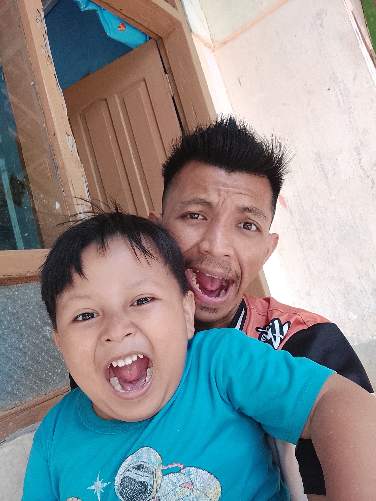 In Kertahayu, Indonesia, two males, one an adult and the other a child, are captured in a close-up selfie. The adult, with spiky dark hair and an open mouth expressing surprise or excitement, is positioned behind the child. The child, with dark hair and a wide, open-mouthed smile, is in the foreground, facing the camera. Both are wearing t-shirts, the child in a bright blue t-shirt with a graphic print and the adult in an orange and black t-shirt. The background shows an interior doorway with a wooden frame, and a textured wall. The lighting appears to be natural and direct, creating strong highlights and shadows.