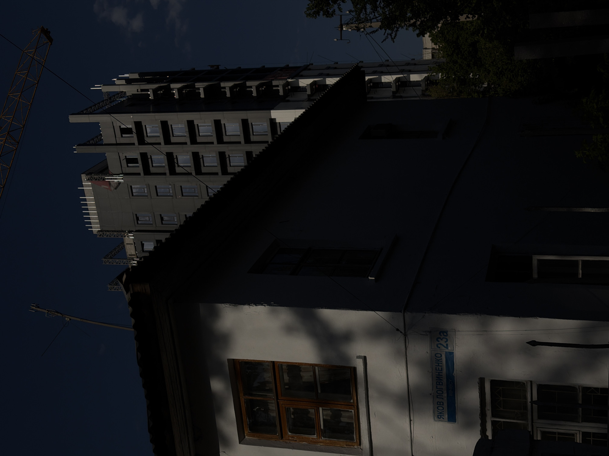 In an urban environment, a low-rise building with a dark, corrugated roof casts a shadow over its front facade, which is painted a light color. A window with wooden frames is visible on the left side of this building, with a second, narrower window to its right. A street sign is mounted on the facade, reading "ЯКОВ ЛОГВИНЕНКО 23a" in blue lettering on a blue background. Behind this building, a tall, unfinished high-rise structure, likely under construction, dominates the background. A yellow construction crane is positioned in the upper right corner of the frame, its boom extending over the scene. The sky is a deep blue, with some scattered clouds. Trees are visible on the right side of the image, and a utility pole with wires is present. The lighting suggests it is either early morning or late afternoon, with strong shadows cast across the scene.