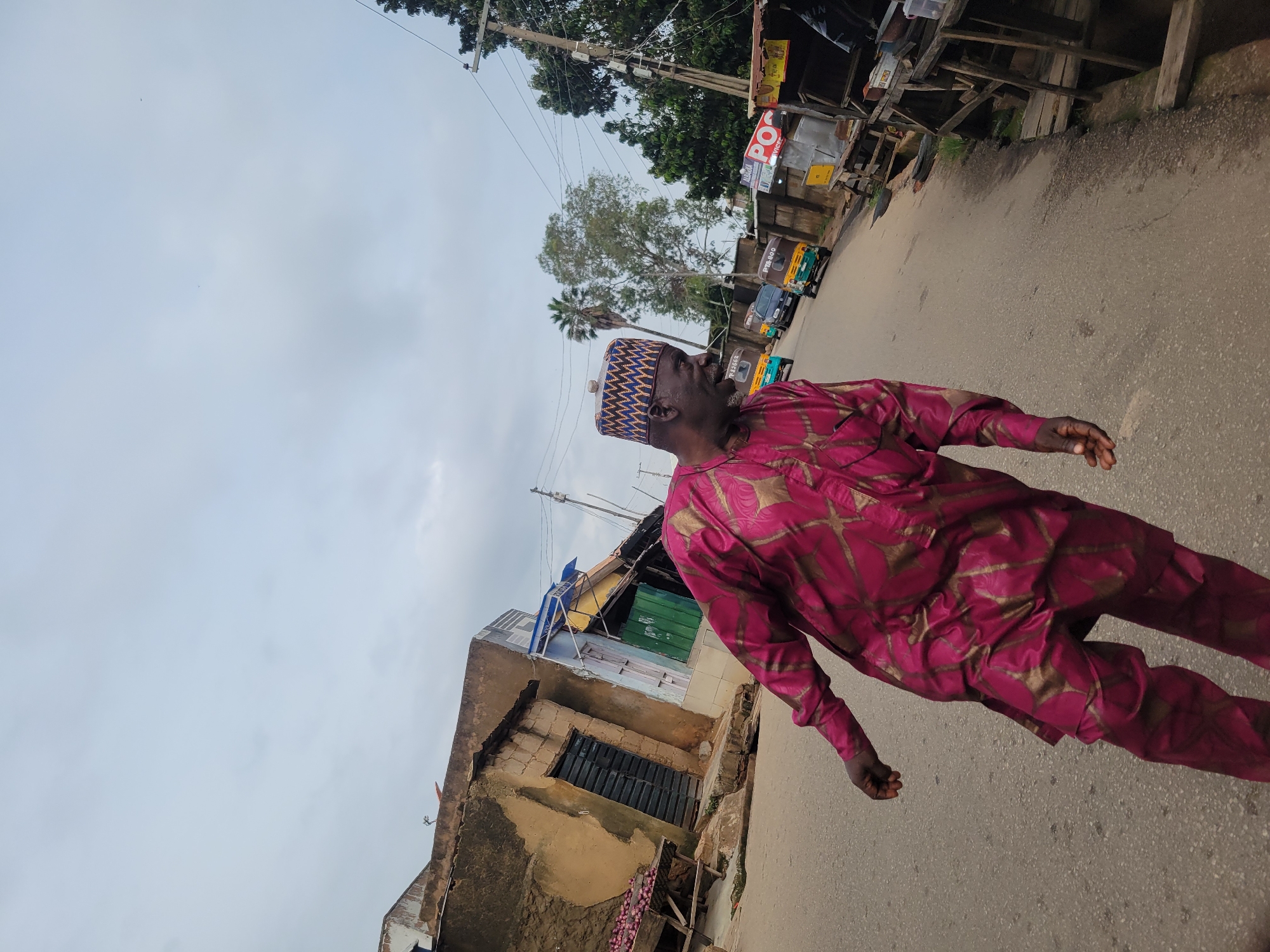 A man wearing a magenta and gold patterned traditional outfit and a patterned cap walks down a street in Jos, Nigeria. The man is facing right and looking slightly upward. The street is paved and lined with buildings and market stalls on both sides. Several vehicles, including auto-rickshaws and a car, are visible on the street. The sky is overcast and appears to be daytime. The overall scene depicts a bustling street in a Nigerian city.