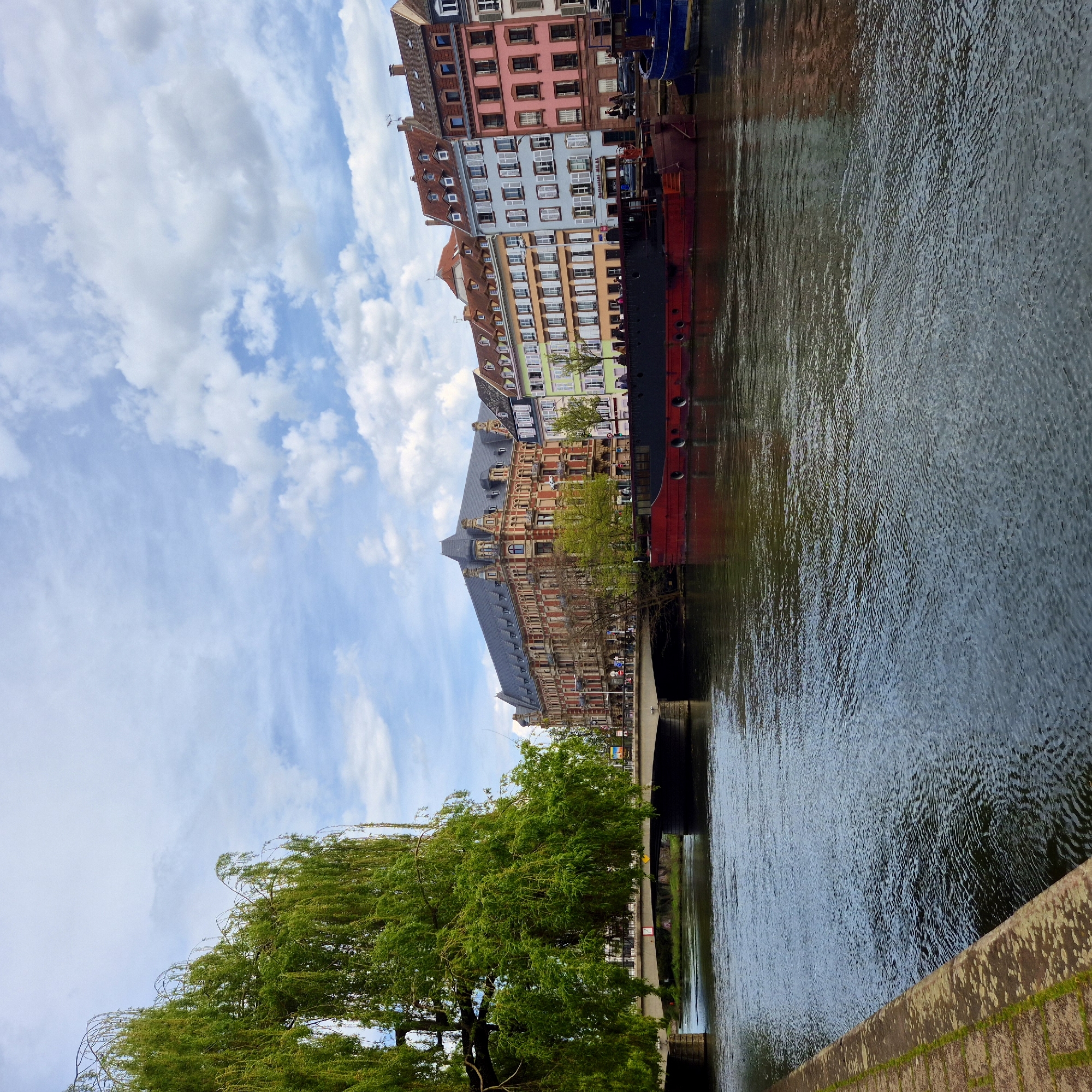 The image captures a canal scene in Strasbourg, France, featuring a waterway flowing adjacent to a row of historic buildings with various architectural styles and colors. A large barge is docked along the canal. A willow tree is positioned on the canal bank. The sky is cloudy. A bridge spans the canal. The photo seems to be taken from a low angle, near water level, on the bank. The water's surface is rippled.