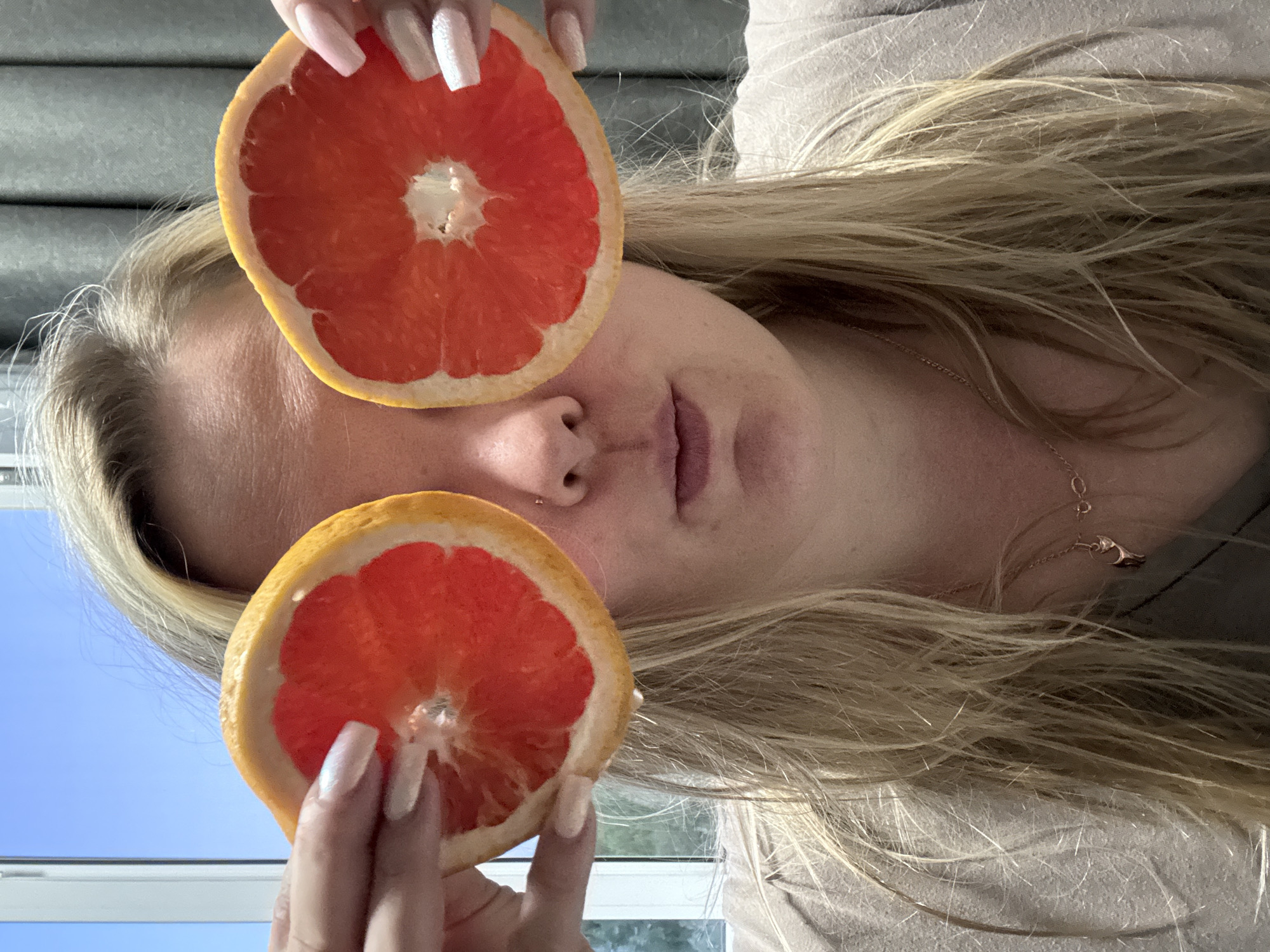 A woman with long blonde hair is holding two halves of a grapefruit in front of her face. The top half is positioned near her forehead, while the bottom half is near her chin. The background includes a window with a view of a blue sky and greenery. The woman's nails are long and painted with a light color. She is wearing a light-colored shirt and a delicate necklace. Марковский, Russia, cannot be determined as the location.