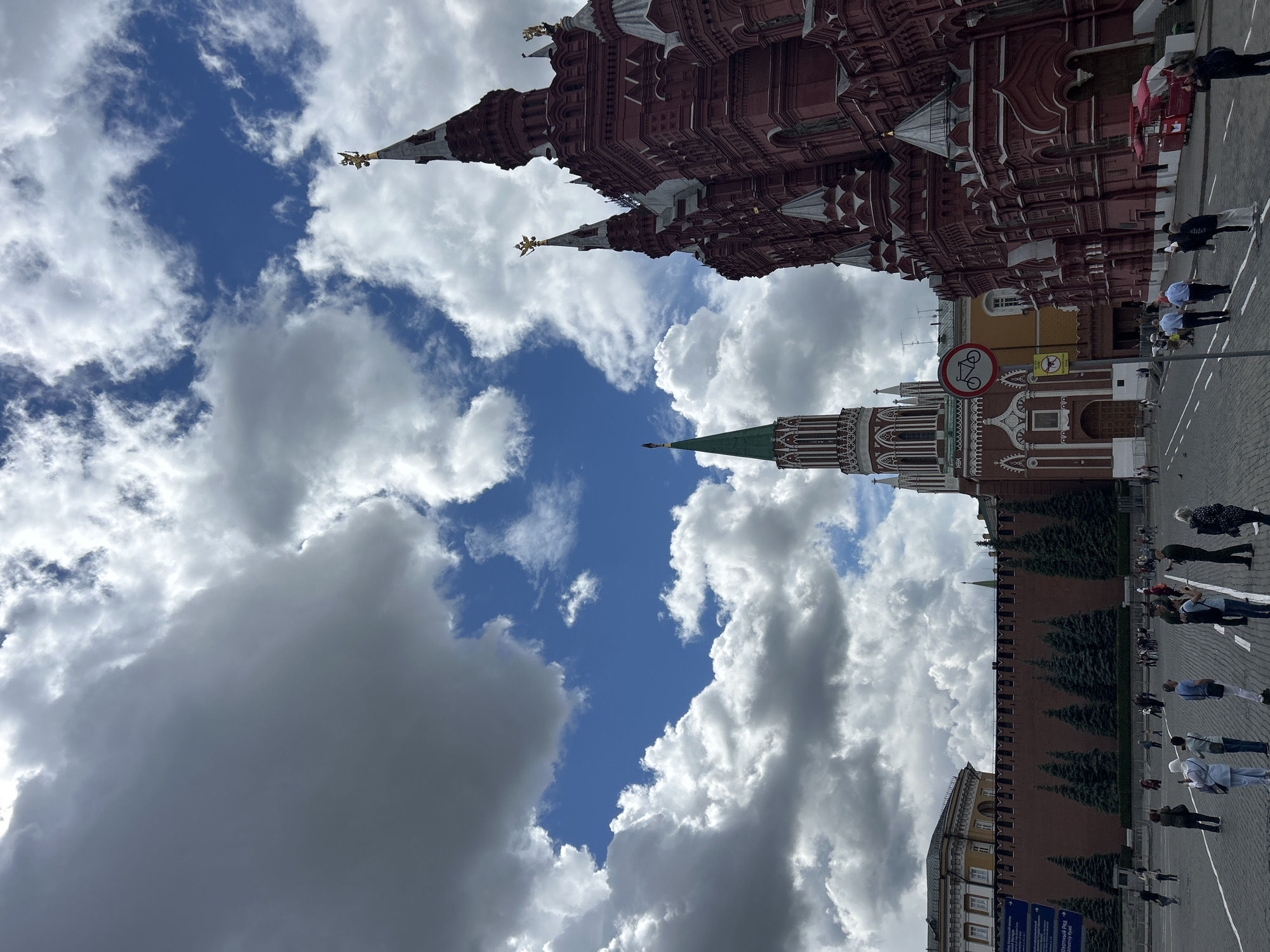 The image depicts the Moscow Kremlin, with a view that includes the walls, towers, and a street scene. The red brick structures of the Kremlin are prominent, featuring intricate architectural details and golden spires against a backdrop of a partly cloudy sky. A sign with a "no bicycles" symbol is visible. People are walking along the street next to the Kremlin wall. The environment suggests a sunny day at a popular tourist destination.