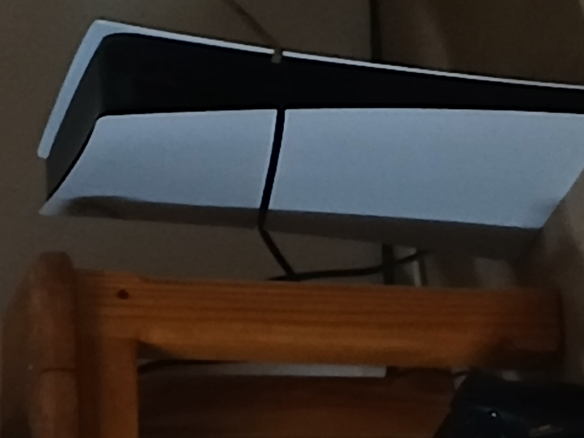The image shows a PlayStation 5 console standing vertically next to a wooden table leg. The console is white and black with a distinctive curved design. A black cable is visible behind the console, connecting it to an unseen power source. The environment appears to be indoors, possibly a living room or gaming setup, with a neutral-colored wall in the background. There is no indication of the city of Dnipro, Ukraine.