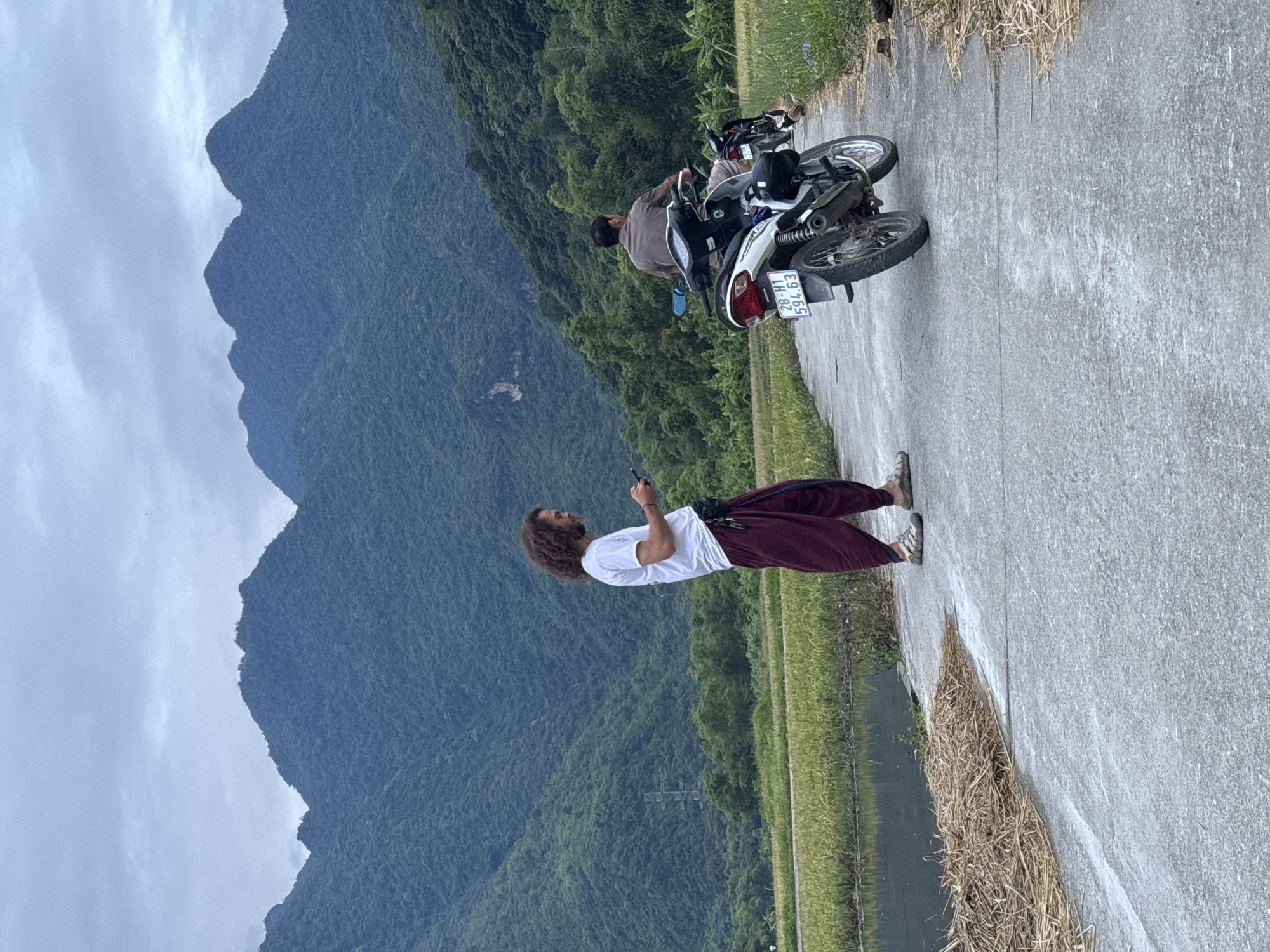 In Chiềng Châu, Vietnam, a person with curly brown hair, wearing a white t-shirt and maroon pants, stands on a concrete road holding a mobile phone. They are looking down at the device, with a black fanny pack around their waist. To the right of the frame, a white motorcycle with a license plate reading "28-H1 594.63" is parked. A person wearing a brown shirt and shorts is seated on the motorcycle. In the background, lush green mountains dominate the landscape under a cloudy sky. To the left of the road, a flooded rice field is visible, reflecting the surrounding greenery. The scene appears to be a rural road in the mountainous region of Chiềng Châu.