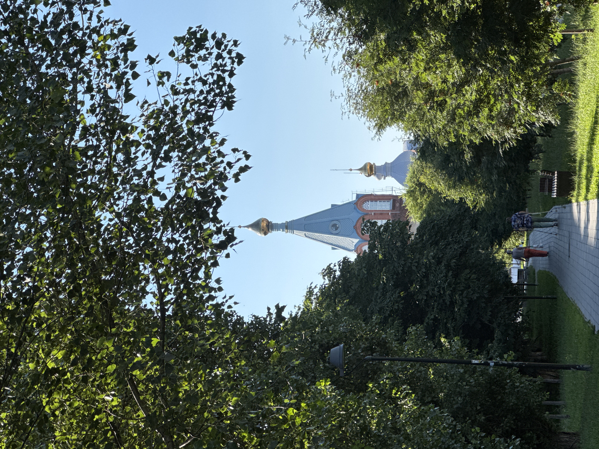 The image depicts a sunlit park pathway bordered by lush green trees. In the background, the iconic golden domes of the Kremlin's Annunciation Cathedral in Nizhny Novgorod, Russia, are visible against a clear blue sky. Two individuals are walking along the paved path, one carrying a white plastic bag and the other using a white cane, suggesting they are either visually impaired or using it for support. The path is illuminated by the sun, casting shadows from the trees. A lamppost stands to the left of the path, and park benches are situated alongside it. The overall atmosphere is peaceful and serene, capturing a moment of daily life in a historic urban setting.