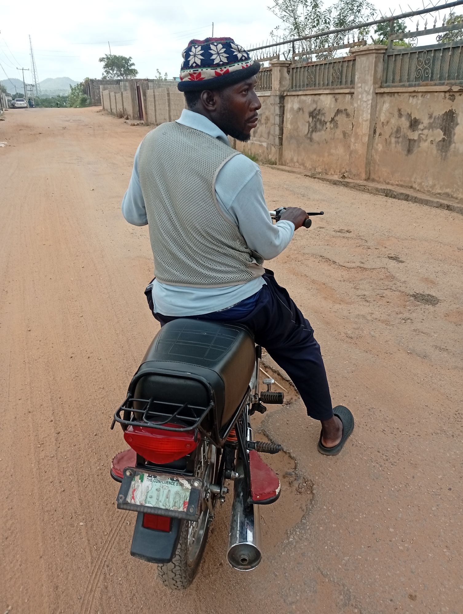 A man is sitting on a motorcycle in Lokoja, Nigeria. The man is Black, wearing a patterned hat, light blue shirt, light grey vest, and dark blue pants. He is looking to the right of the frame. The motorcycle is red and black. The motorcycle has a license plate that reads "THE CONFLUENCE STATE." The road is dirt and has some potholes. There are buildings and fences in the background. The sky is overcast.