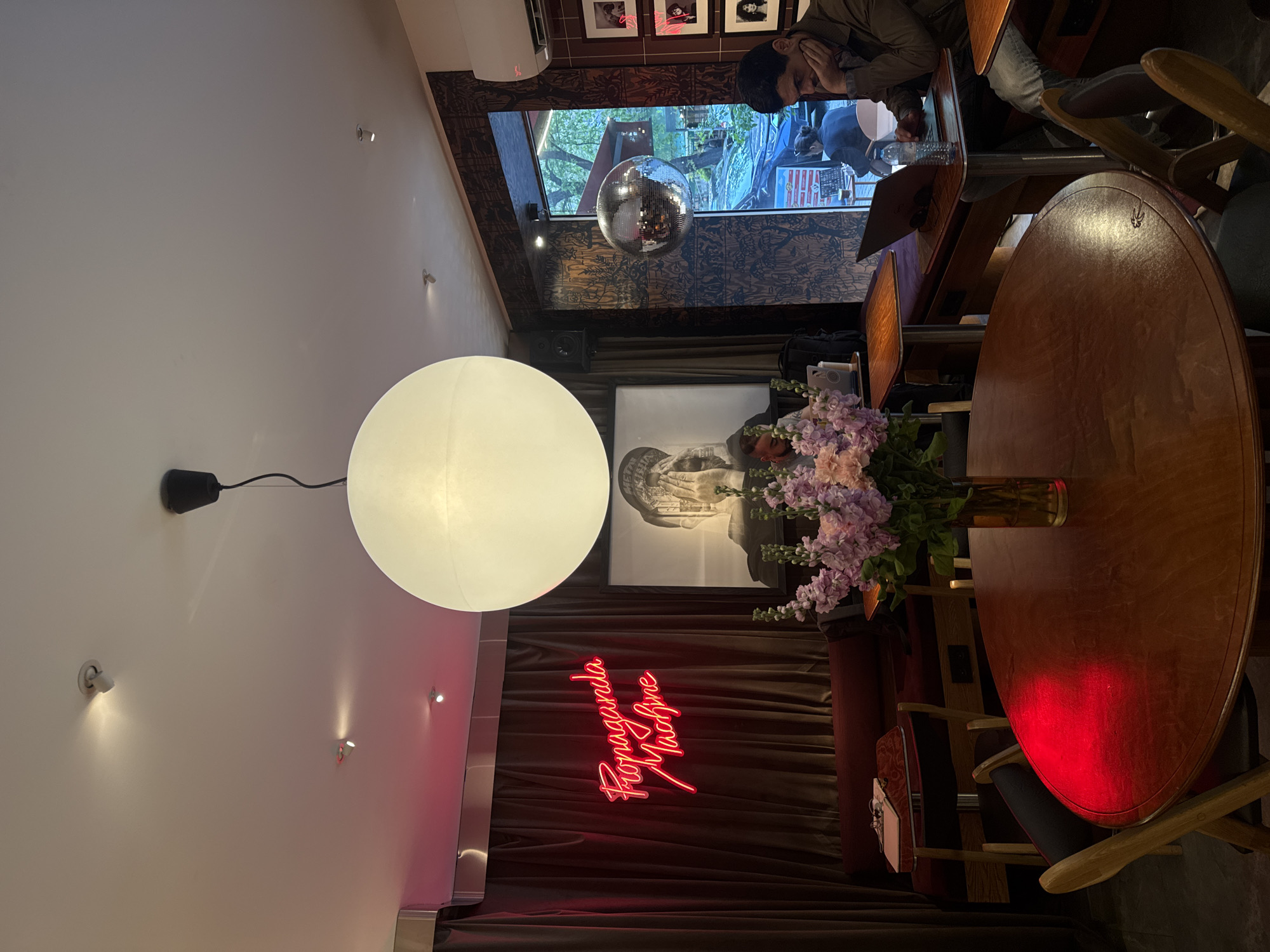 This rotated image captures the interior of an atmospheric establishment, likely a cafe or bar, in Yerevan, Armenia, characterized by warm, dim lighting and eclectic decor.
On the left side of the frame, a large, glowing white spherical pendant light hangs from the ceiling, casting a soft, diffused illumination. Below this, against a plain wall and dark, plush curtains, a vibrant red neon sign prominently displays the text "Propaganda Machine" in a cursive font. Small, directional spotlights are also visible on the wall sections.
Towards the center, a round wooden table serves as a focal point, holding a tall vase filled with a lush bouquet of purple and pink flowers. Behind this table, a framed black and white artwork depicting a classical bust adds to the artistic flair.
The background features a large window offering a glimpse of an outdoor scene with trees, suggesting it is evening or nighttime. A glittering disco ball hangs within the window frame, reflecting light and enhancing the festive atmosphere. Below the window, the wall is adorned with a dark, patterned wallpaper.
In the upper right corner, a man is seated at a wooden counter or table, leaning forward with his hand resting near his face, appearing thoughtful or engaged. Other indistinct figures can be seen in the background or reflections, indicating patronage. The overall scene suggests a stylish, cozy, and vibrant space for socializing or dining.