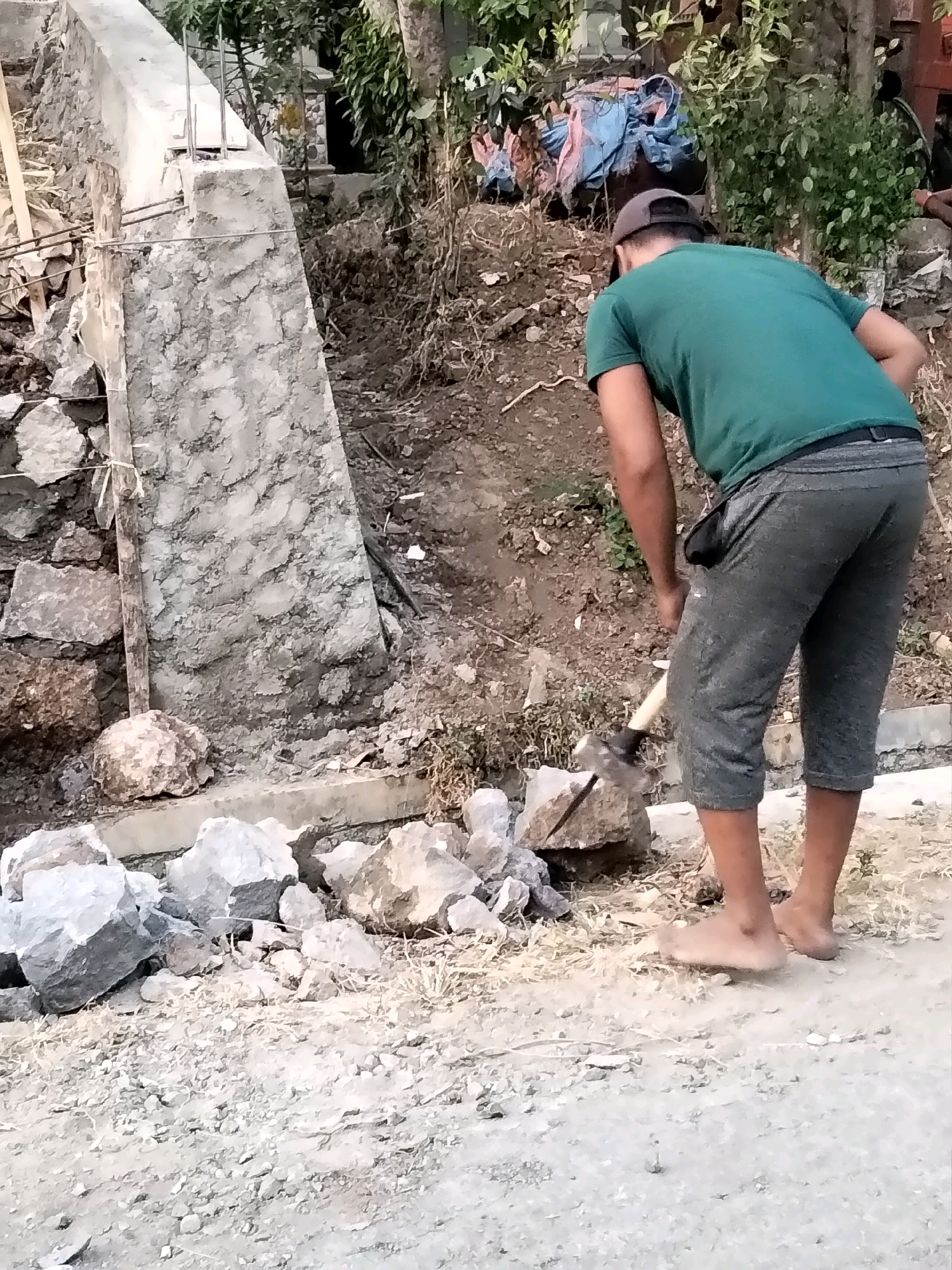 A person is seen from behind, wearing a green t-shirt and grey capri pants, working on breaking rocks with a sledgehammer. The person is barefoot. The scene appears to be outdoors, with a construction site in the background, featuring a concrete structure and piles of rocks. The ground is uneven and covered with gravel and dry vegetation. The environment suggests a rural or semi-urban area. The image lacks specific details to pinpoint the exact location within Jenangan, Indonesia, other than it being a potentially ongoing construction project.