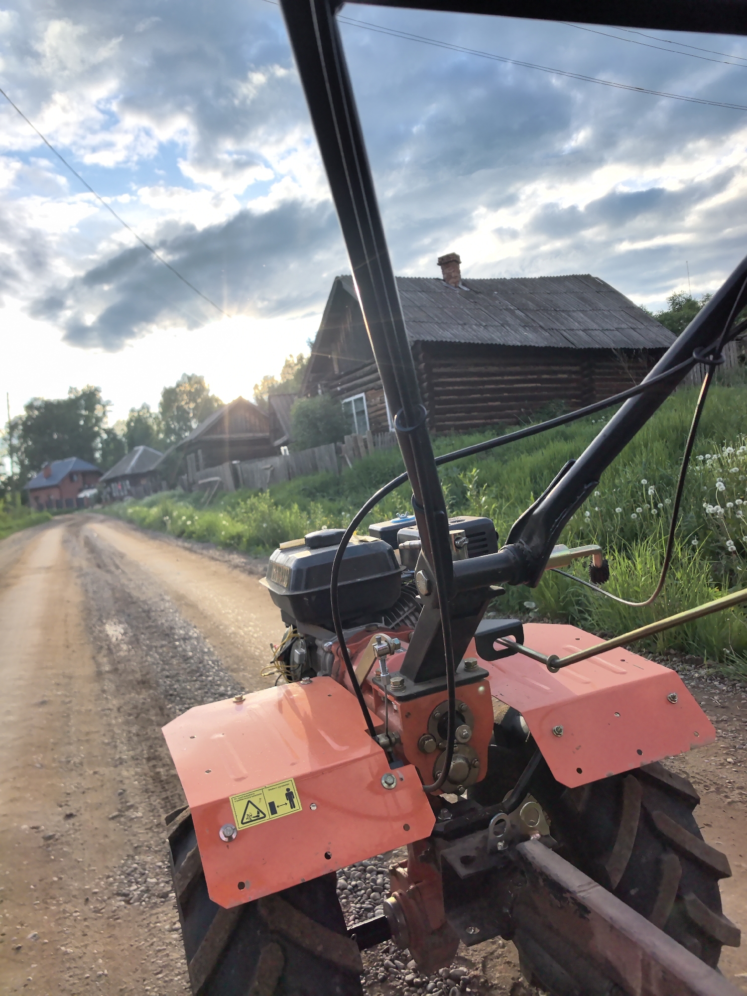 A medium shot shows a gravel road and surrounding rural environment from the perspective of an agricultural tiller in Unknown, Unknown. The tiller's rear features orange mudguards, black tires, and a central engine. The tiller's handles, also black, frame the composition, partially obscuring the background. The road stretches into the distance, flanked by wooden houses and fences. Overgrown grass and wildflowers border the road. Overcast sky with a setting sun. The sun is low on the horizon, partially obscured by clouds. Power lines stretch across the sky above the road.