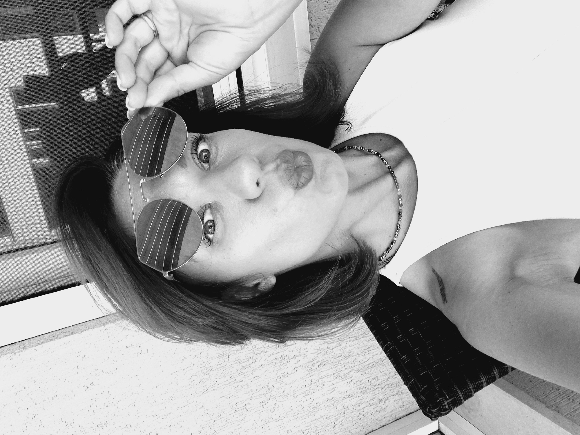 A black and white, medium shot, selfie of a woman sitting on a woven chair outdoors. The woman is wearing sunglasses and a white tank top. She is holding her sunglasses up with her right hand and puckering her lips. She has a beaded necklace around her neck and a tattoo on her left shoulder. The background is a textured wall and a window screen.