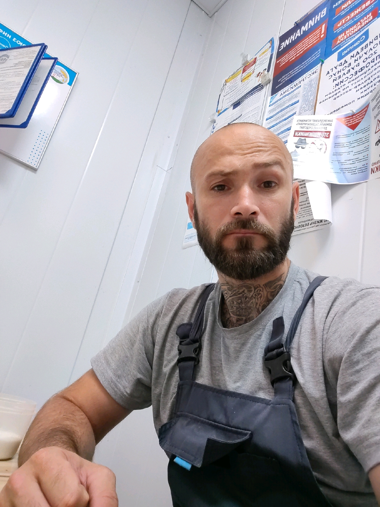 A man with a shaved head, beard, and neck tattoos is sitting in front of a white paneled wall. He is wearing a grey t-shirt and dark blue overalls. Several posters are pinned to the wall behind him, some with Russian text. To the left of the man, a blue folder with documents is attached to the wall. In the foreground, his hand is resting on a wooden surface, and to his left, a white plastic container is visible. The overall setting appears to be an indoor workspace, possibly in Pinsk, Belarus, given the context of the posters.