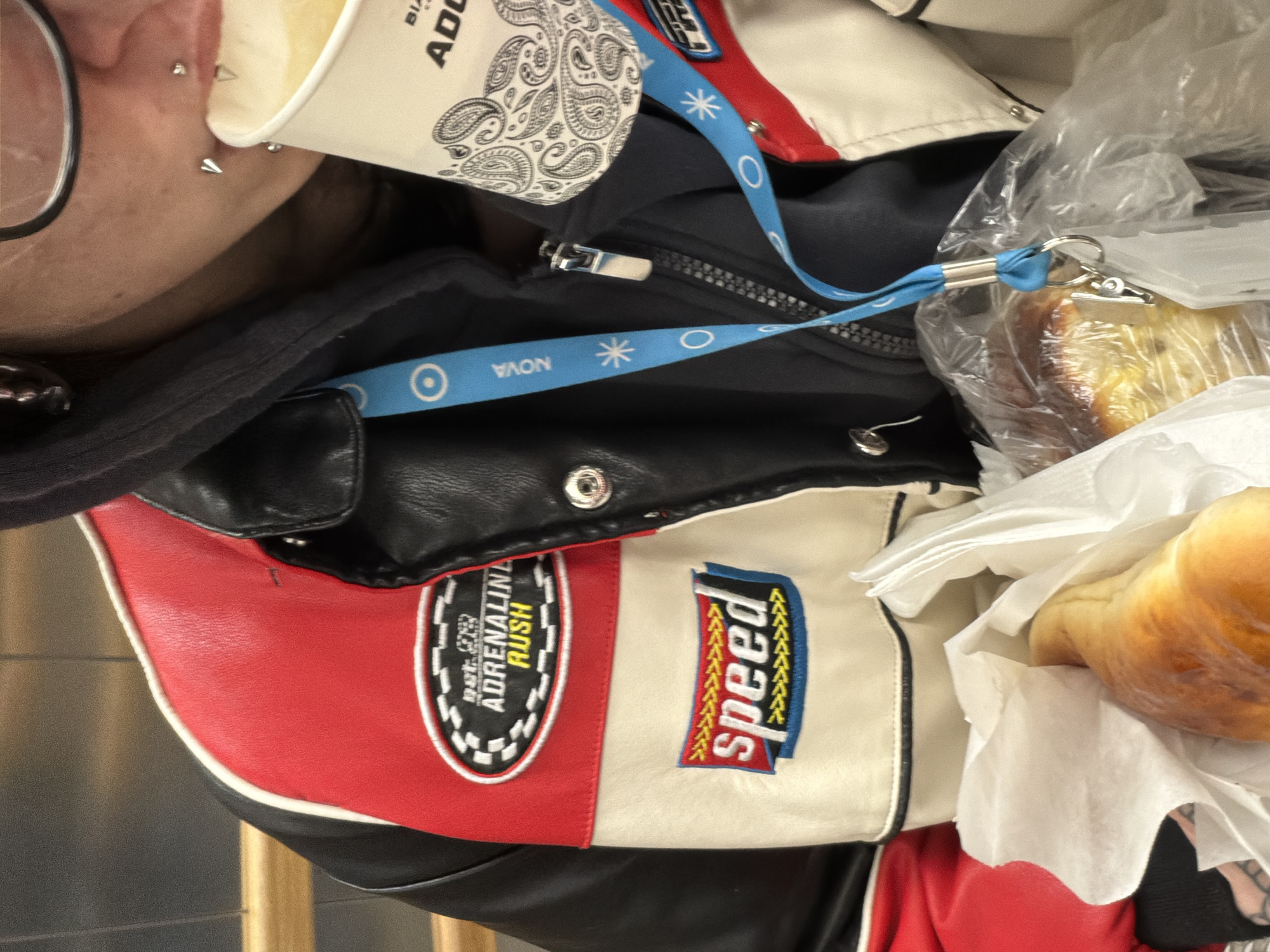 The image shows a close-up, overhead view of several items. A person's face with glasses and multiple ear piercings is visible on the left edge. Next to the person is a paper cup with a paisley pattern and the partially visible word "ADO." Below the cup is a black hooded garment with a zipper. At the bottom of the image is a red and white jacket with black accents and patches. The jacket has patches featuring the words "ADRENALINE RUSH" and "speed" with stylized fonts. Lying on top of the jacket is a wrapped bread roll in a clear plastic bag and white napkin. A blue lanyard with the word "NOVA" and snowflake designs is visible near the jacket's collar. The location in the city of Sofia, Bulgaria, cannot be determined from the image.