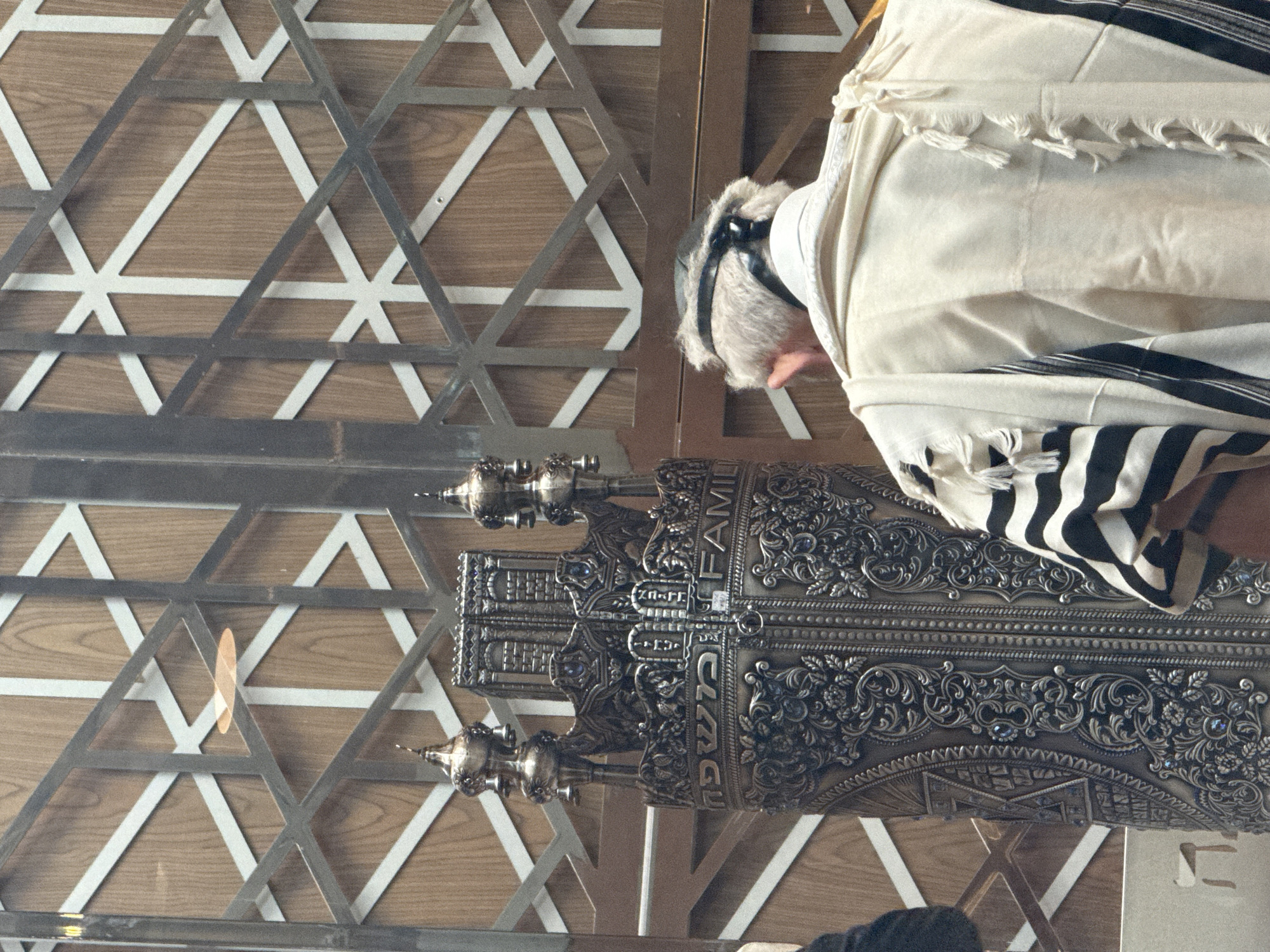 The image captures a poignant moment within a Jewish religious setting, likely a synagogue or communal space in Mexico City. The scene is presented from a slightly elevated, over-the-shoulder perspective of an individual engaged in prayer or religious observance.
The main subject is a person, seen from behind, with their head respectfully bowed. They are adorned in a traditional white tallit (Jewish prayer shawl) featuring prominent black stripes and visible fringes (tzitzit). A head covering, possibly a kippah or yarmulke, is visible beneath the tallit.
Central to the foreground is a magnificently ornate Sefer Torah (Torah scroll), encased in an elaborate, decorative metal covering. This casing, appearing to be crafted from silver or bronze-toned metal, is richly adorned with intricate filigree, floral motifs, and what appear to be small, sparkling blue or purple stones. Two decorative finials (Rimonim) crown the top of the Torah case. Clearly visible on the casing are the English word "FAMILY" and the Hebrew word "משפחה" (mishpacha), both meaning family.
The background features a wall composed of wooden panels, overlaid with a distinctive geometric metal screen. This screen is characterized by interlocking triangular patterns, subtly incorporating the motif of the Star of David, which reinforces the spiritual context.
The lighting is soft and consistent, indicating an indoor environment. The image evokes a sense of deep reverence and tradition, capturing a moment of devotion during a religious service or personal contemplation.