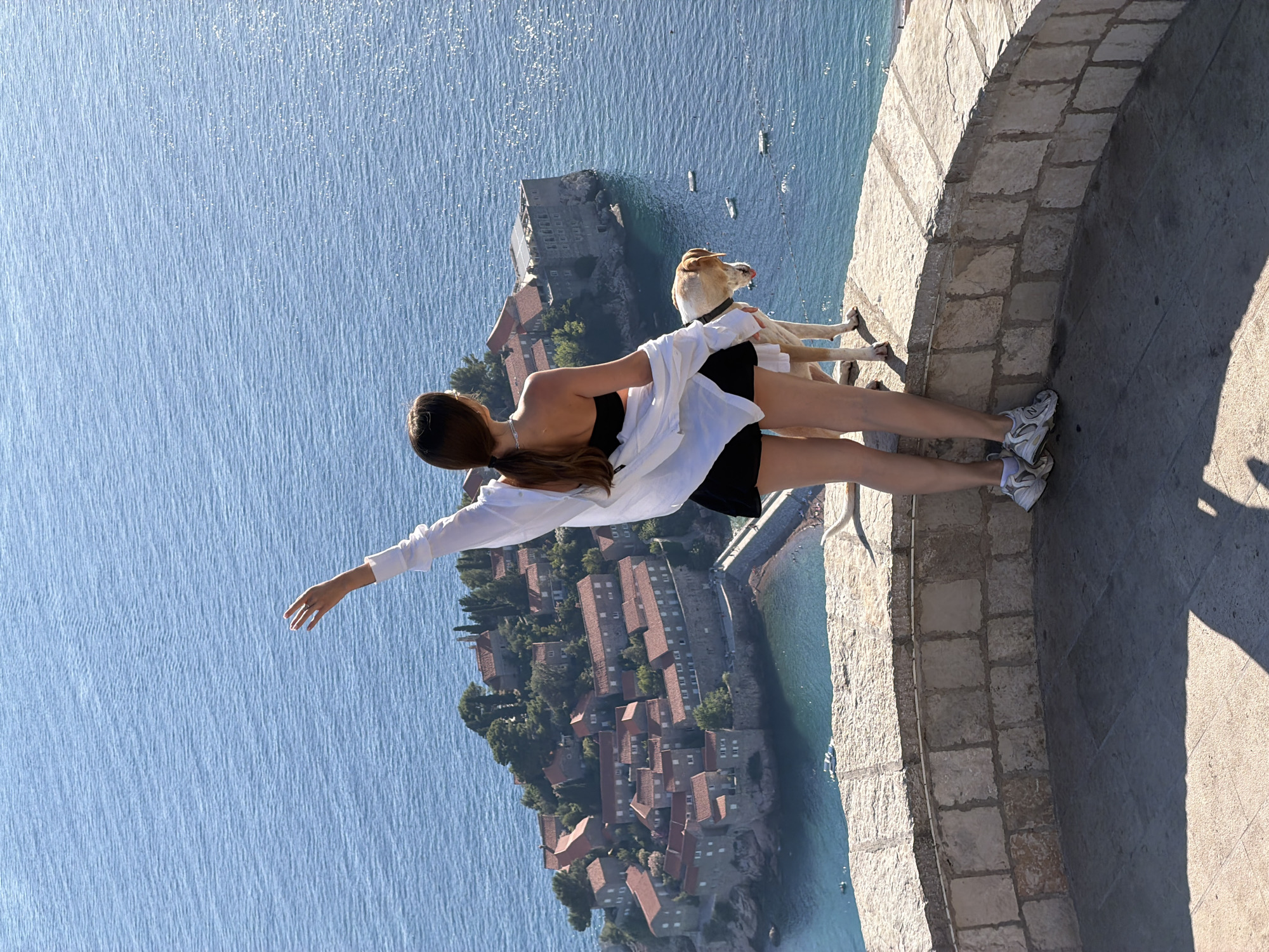 A young woman and a light-colored dog stand on a stone balcony overlooking the island of Sveti Stefan in Montenegro. The woman, with her back to the camera, wears a white unbuttoned shirt over a black strapless top and black shorts. Her left arm is raised, with her hand in a wave gesture. The dog, a light brown and white Labrador mix, stands next to her, looking out at the sea. The island of Sveti Stefan is visible in the background, a cluster of red-roofed buildings with stone walls, connected to the mainland by a narrow causeway. The sea is a deep blue, with sunlight glinting off its surface. The sky is clear and blue.