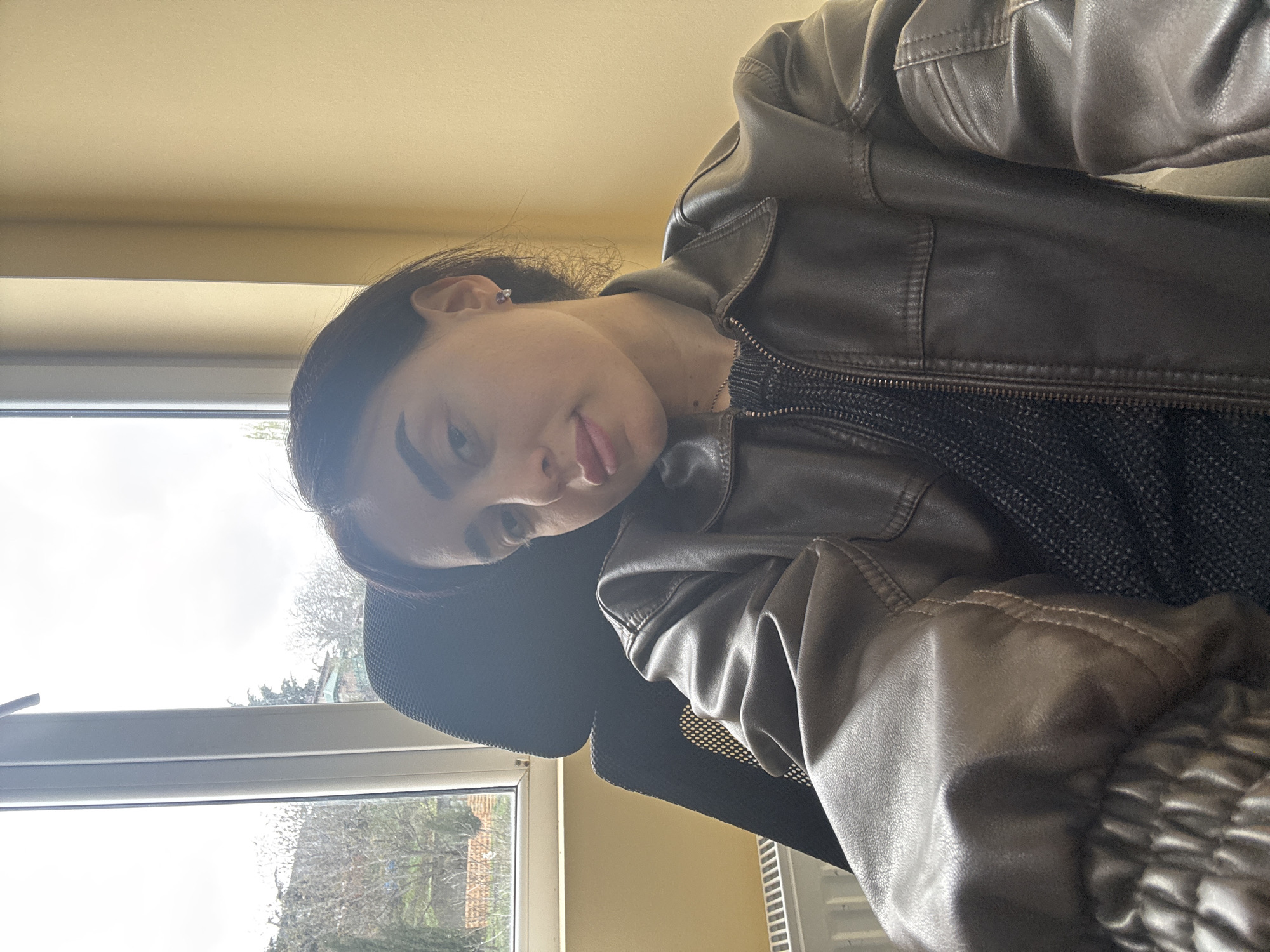 A woman with dark hair is posing in what appears to be an indoor setting in front of a window. She is wearing a brown leather jacket over a dark-colored knitted top. The window offers a view of trees and a building exterior, possibly indicating a residential area. The location cannot be determined to be Tbilisi, Georgia with the details provided.