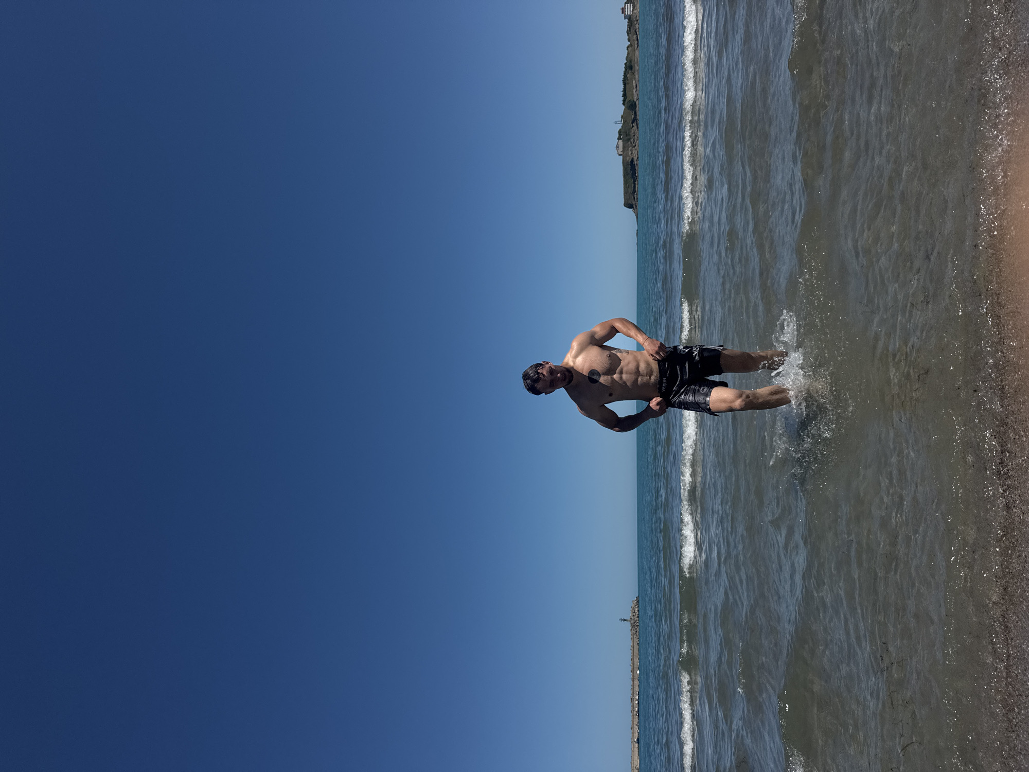 The image depicts a man standing in the shallow water of a beach, with waves gently lapping around his legs. He is shirtless, wearing black swim shorts, and his tanned skin is glistening in the sunlight. The man is looking towards the viewer with a slight smile, his pose suggesting a playful or energetic mood.
The scene is set at a beach with clear blue skies above. The water is a clear, greenish-blue, with small waves breaking and creating a foamy white crest. In the background, a long pier or breakwater stretches into the sea, with a small structure resembling a lighthouse or beacon at its end. The coastline is visible in the distance, appearing as a strip of land with some vegetation and structures. The beach itself has a pebbly texture, with small shells and debris scattered along the waterline. The overall impression is of a bright, sunny day, likely during the summer months, with the setting being a coastal town.
The image appears to have been taken during daylight hours, as indicated by the bright sunlight and clear visibility. The sky is a deep, vibrant blue, suggesting fair weather. The location context indicates Tsarevo, Bulgaria, which is a coastal town known for its beaches and seaside activities. The man's attire and the beach setting strongly imply a leisure activity, such as swimming or enjoying a day at the beach. There is some visible text on the man's swim shorts, but it is too small and blurry to be legible.