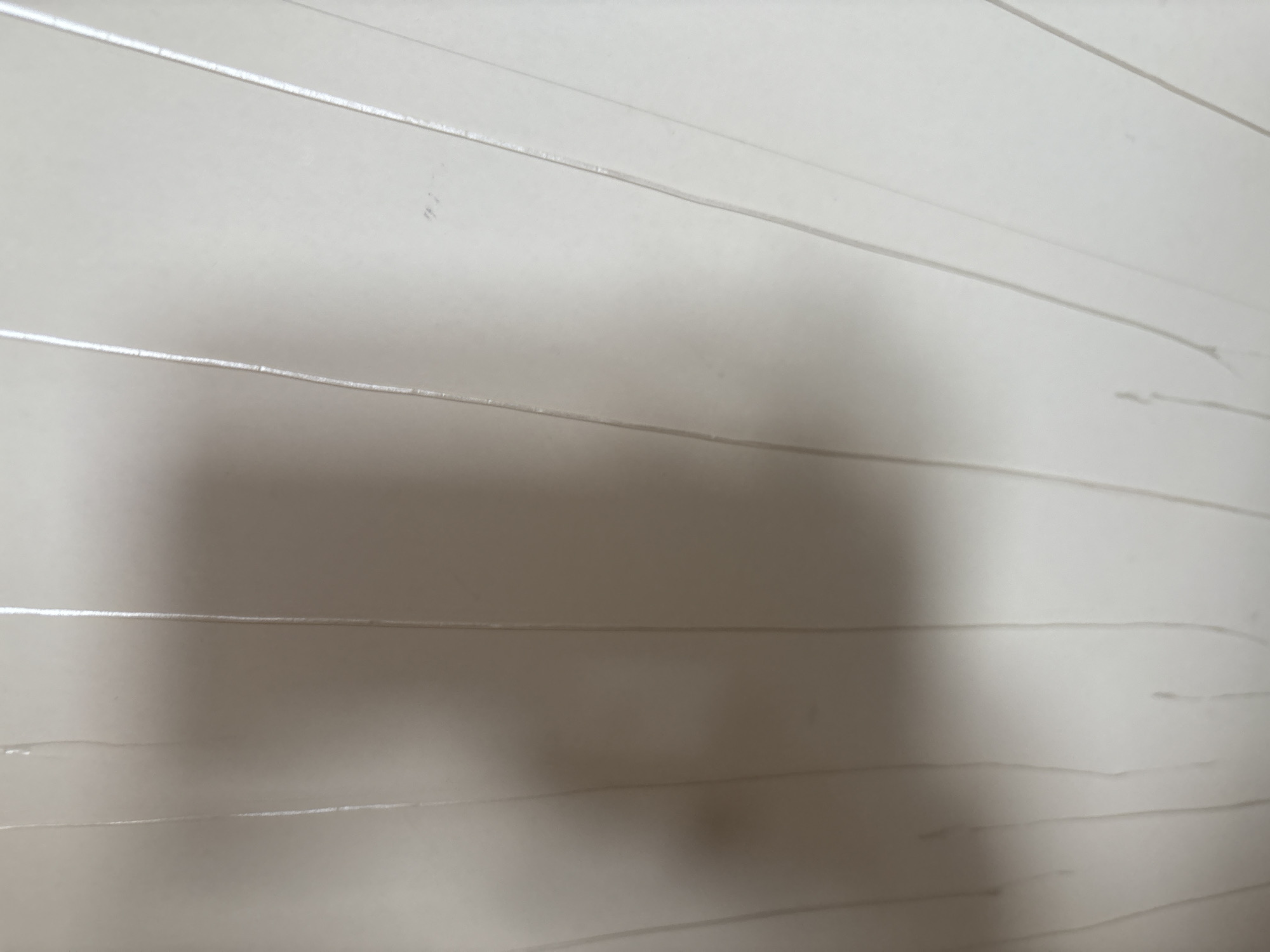 The image shows a close-up of a light-colored wall with vertical lines. There is a shadow on the left side of the wall, suggesting the presence of an object or person out of frame. The wall appears to be made of paneling or has a textured surface. The lighting is dim, creating soft shadows and highlights. The scene is devoid of any specific identifiable features that would allow for its location to be determined in the city of Orekhovka, Russia.