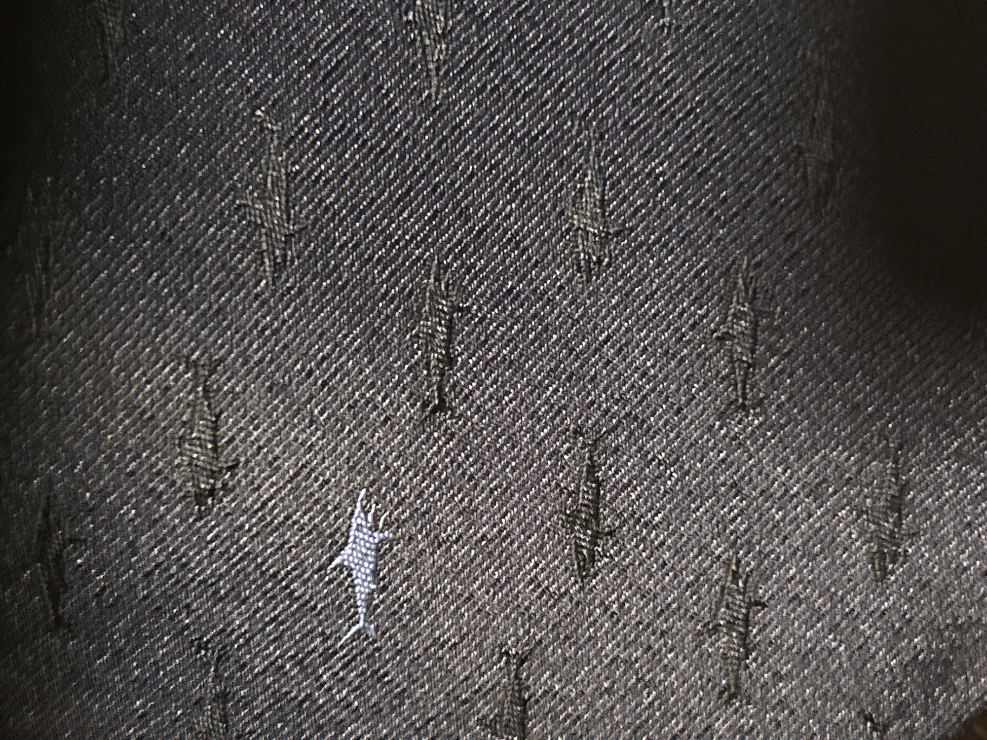 The image is a close-up, top-down view of dark blue denim fabric with a subtle textured weave. Scattered across the fabric are small, light-colored embroidered fish shapes. One fish, more prominently embroidered and appearing to be a marlin, is located in the upper left quadrant of the image. The rest of the fish are smaller and less distinct, appearing as dark gray or black silhouettes against the denim. The fabric exhibits some wear and tear, with areas of lighter threads and small holes visible, particularly in the lower half of the image. There is a gradual gradient of darkness from the bottom of the image towards the top, suggesting uneven lighting or a shadowy area at the bottom. The scene is not located in Moscow, Russia.