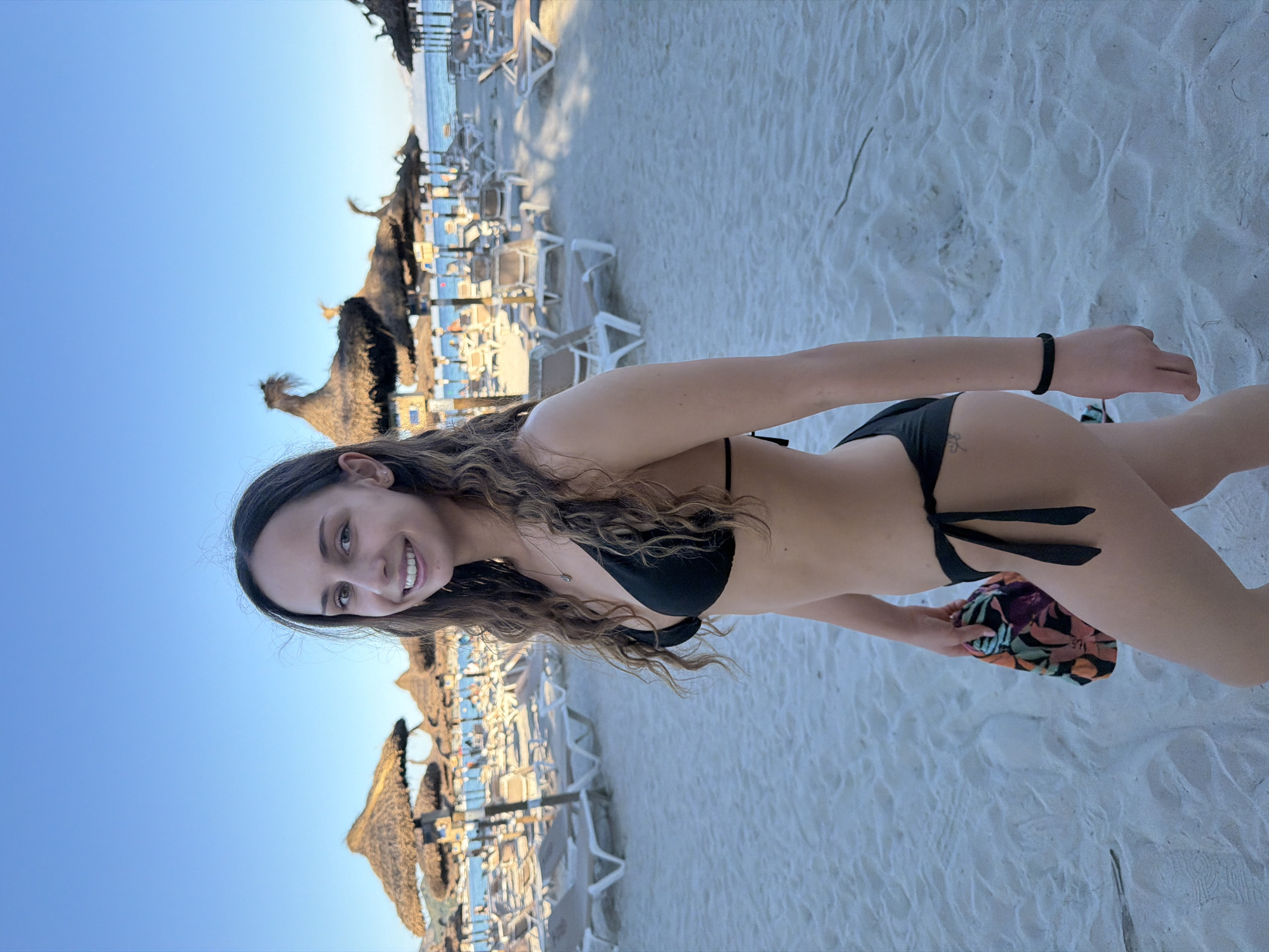 A woman in a black bikini poses on a white sandy beach in Alcúdia, Spain. She is smiling and holding a colorful pouch. In the background, there are straw umbrellas and white beach chairs. The sky is blue.