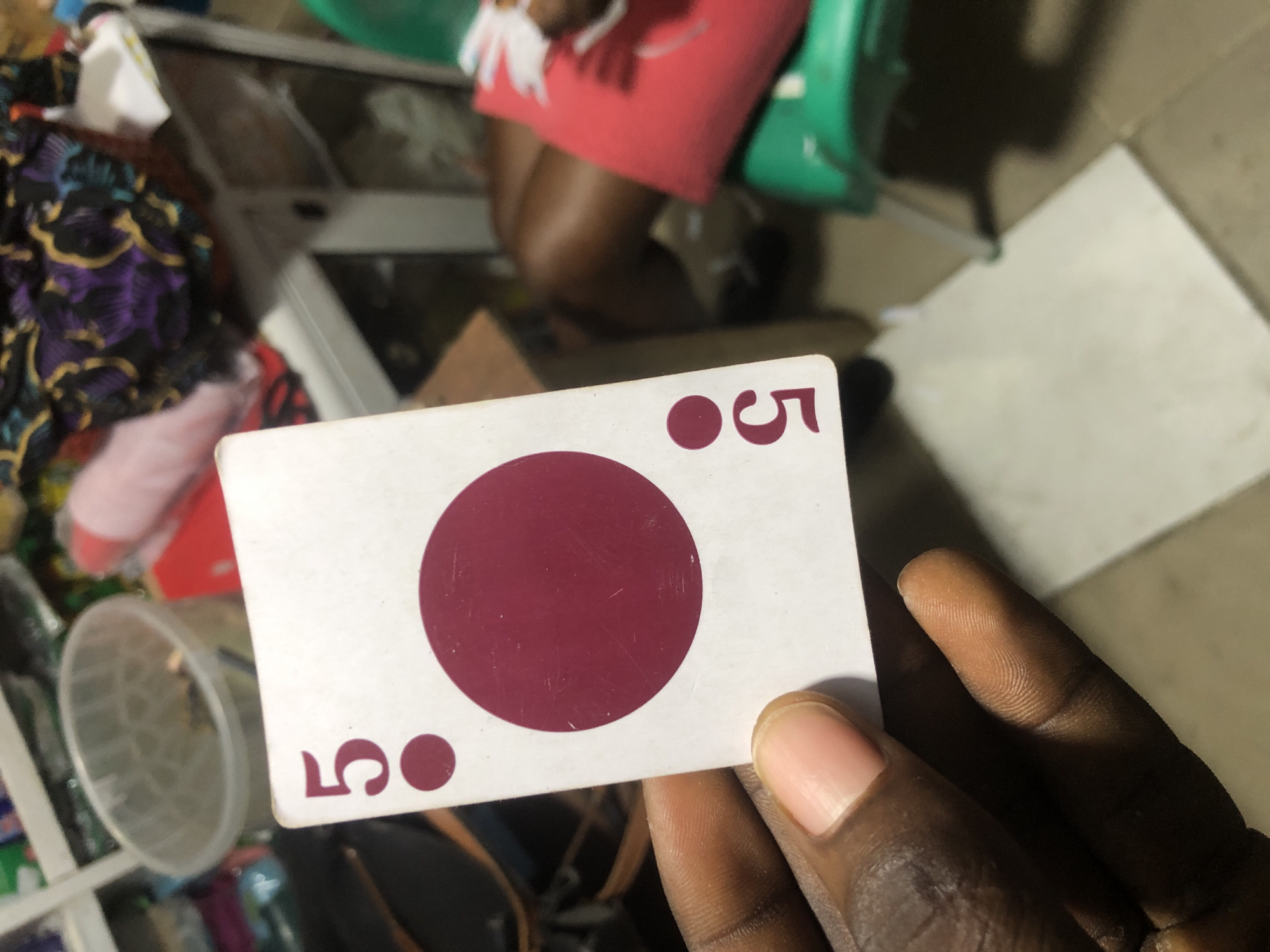 A hand holds up a playing card, the "five of diamonds", which is white with maroon pips. The card is held in the foreground, obscuring much of the background which is blurry and appears to be an indoor market or shop. In the background, there is a clear plastic tub, various colorful fabrics, and a green plastic chair. The scene is likely Ado, Nigeria, based on the provided context.