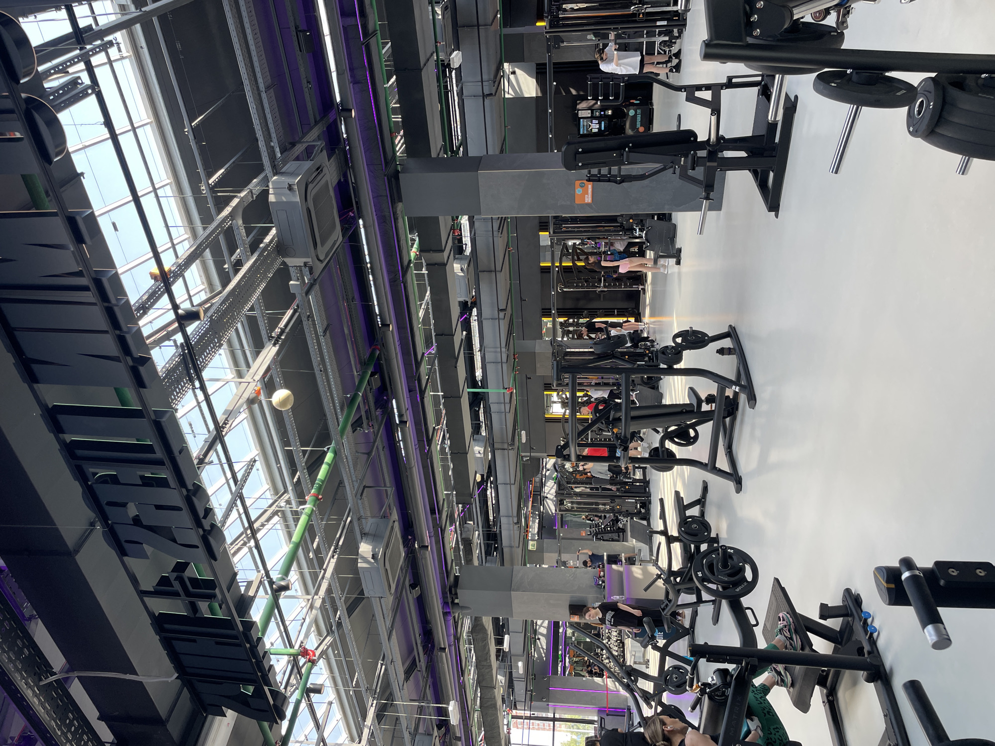 The image depicts the interior of a large, modern gym located in Kazan, Russia. The gym has a high ceiling with a skylight providing natural light. Various weightlifting machines are arranged throughout the space, with several individuals using the equipment. The gym features prominent signage. Metal beams, ductwork, and pipes are visible in the ceiling structure. The color scheme incorporates dark grey and purple accents.
