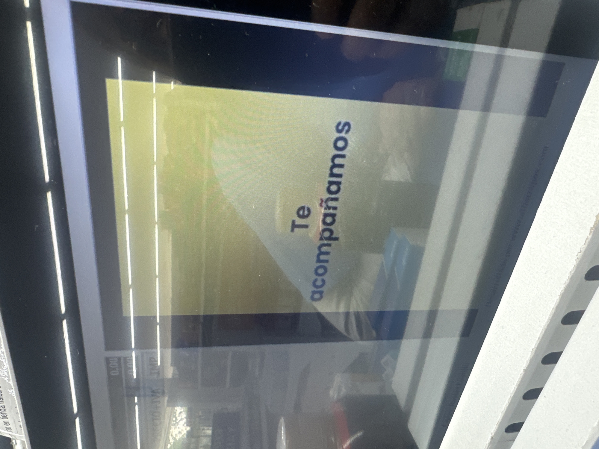 The image shows a close-up of a screen displaying the Spanish phrase "Te acompañamos," which translates to "We accompany you." The screen appears to be part of a point-of-sale system, as indicated by the "0.00" amount visible in the lower-left corner. The reflection on the screen suggests an outdoor setting, and there is a reference to "tienda física" (physical store) in the blurry text at the bottom left. Additionally, a white, possibly metal, frame with rounded holes is visible on the right side of the screen, suggesting the kiosk is placed outside. There is no information about people or actions in the image. The location in the city of Parroquia Ocumare, Venezuela, can not be confirmed.