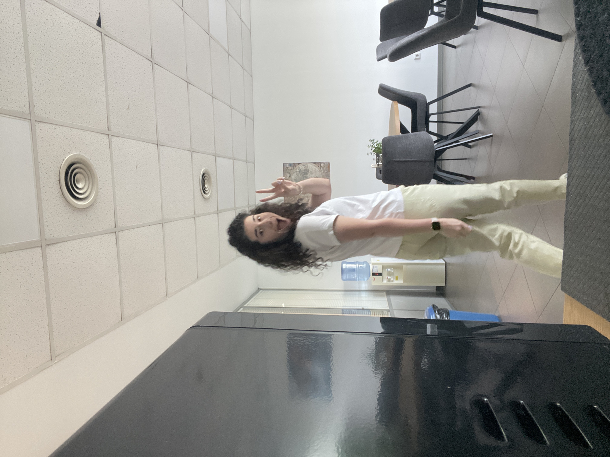 A person with long, dark, curly hair, wearing a white t-shirt and light-colored pants, is making a peace sign with their left hand. They are standing in what appears to be an office setting. The environment features a suspended ceiling with round ventilation ducts, office chairs, a water cooler, a large black object (possibly a refrigerator or cabinet) in the foreground, and a picture on the wall. I cannot determine the location is Sofia, Bulgaria based on the image alone.