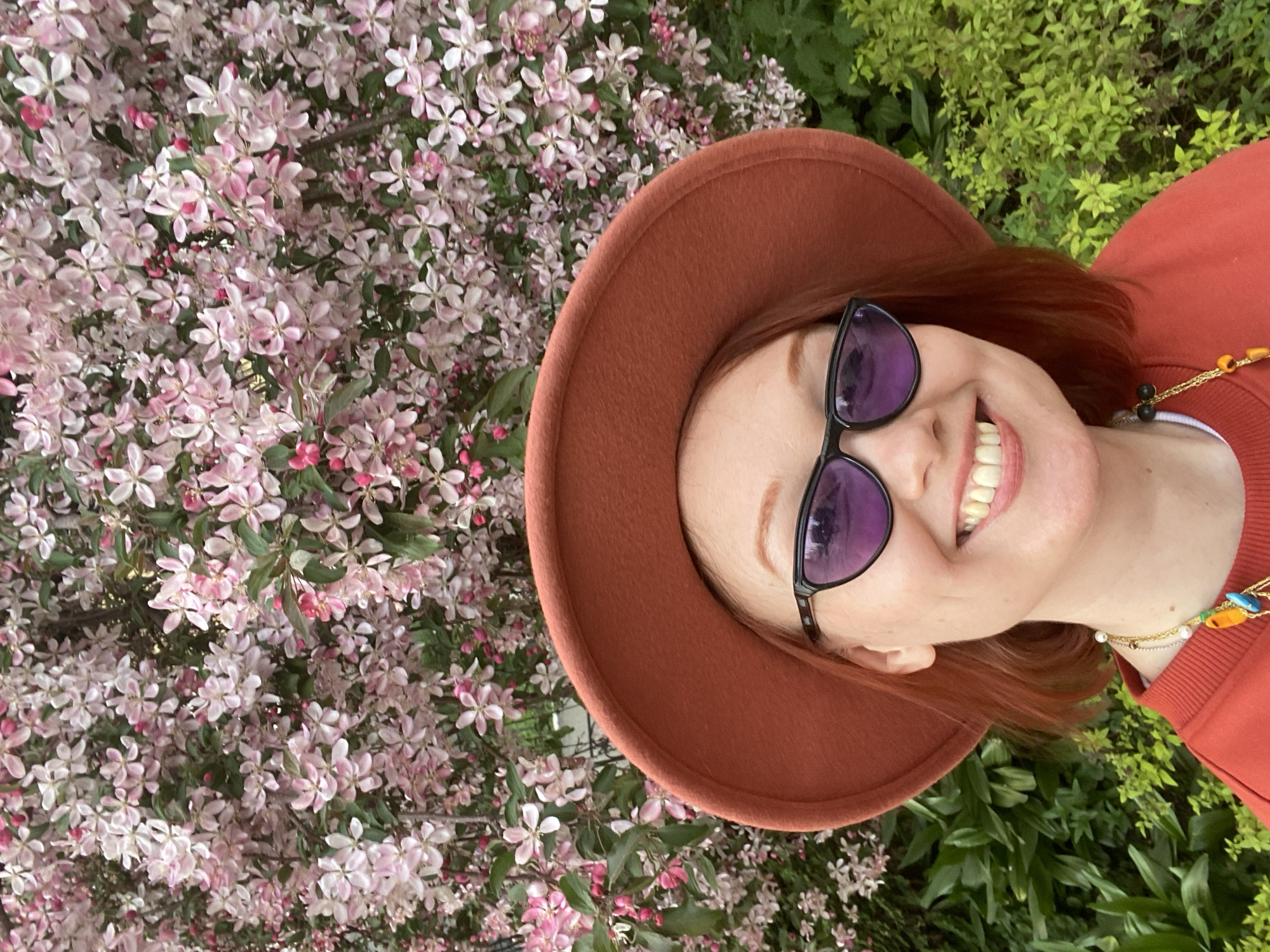 A woman with auburn hair, wearing a brown hat and sunglasses, smiles broadly at the camera. She is surrounded by flowering branches with small pink and white blossoms. The background also includes green foliage. The image was taken outdoors in daylight. The location cannot be determined to be Yekaterinburg based on the visual data.
