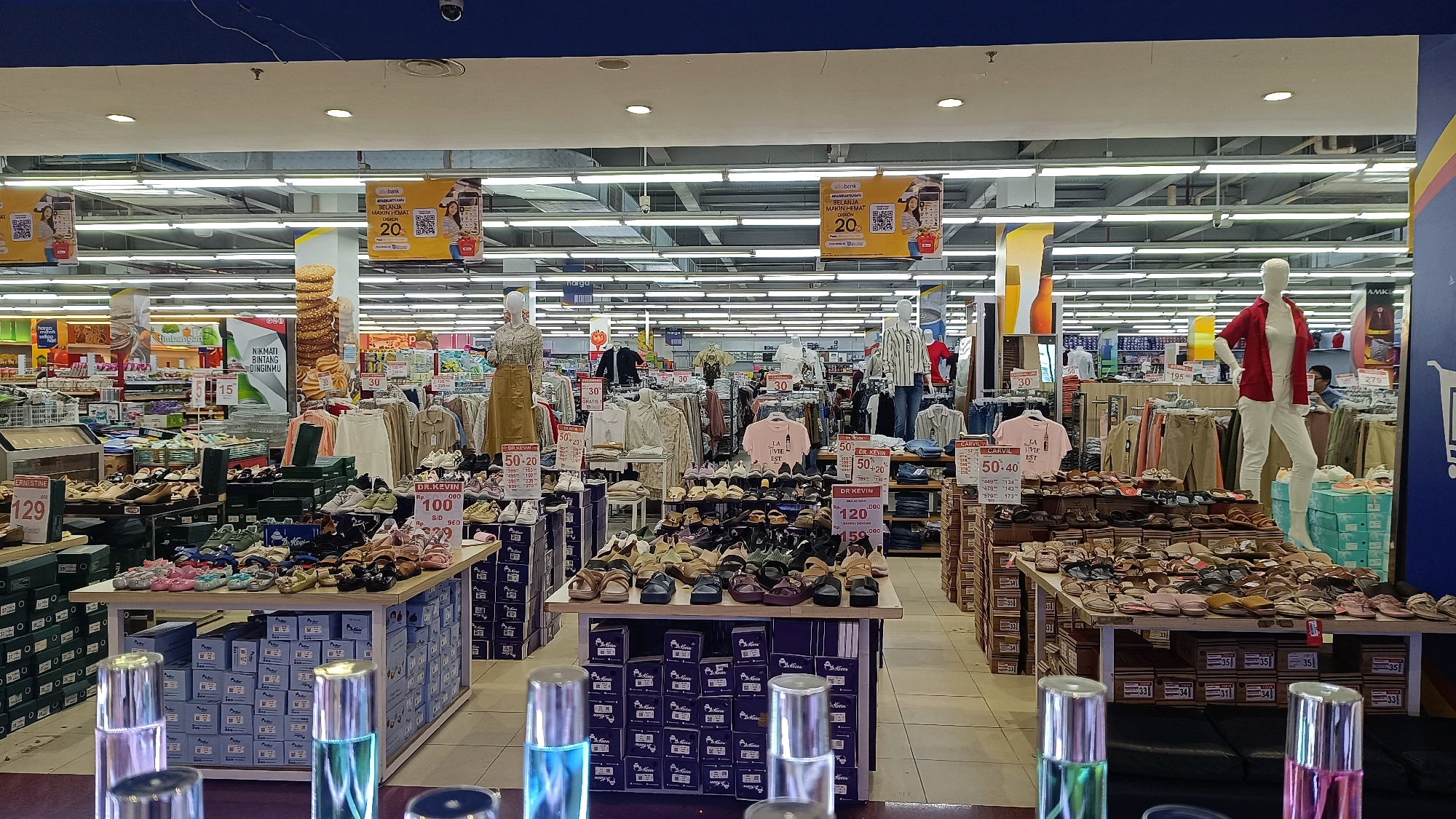 The image depicts a retail store, likely a department store or supermarket, in Tomohon, Indonesia. The environment is brightly lit with fluorescent ceiling lights. The foreground shows a display of various footwear, including sandals and shoes, arranged on tables and stacked in boxes. Some of the boxes have price tags and labels. Two mannequins are visible, one wearing a floral-patterned top and a long skirt, and another in a red jacket and white pants, both positioned amidst clothing displays. Various racks of clothing, including shirts, pants, and dresses, are arranged throughout the store. Promotional banners and signs with discounts and brand names are hanging from the ceiling and on display racks. In the background, aisles of shelves stocked with products are visible, suggesting a large retail space. The overall scene captures a typical retail environment with a focus on apparel and footwear.