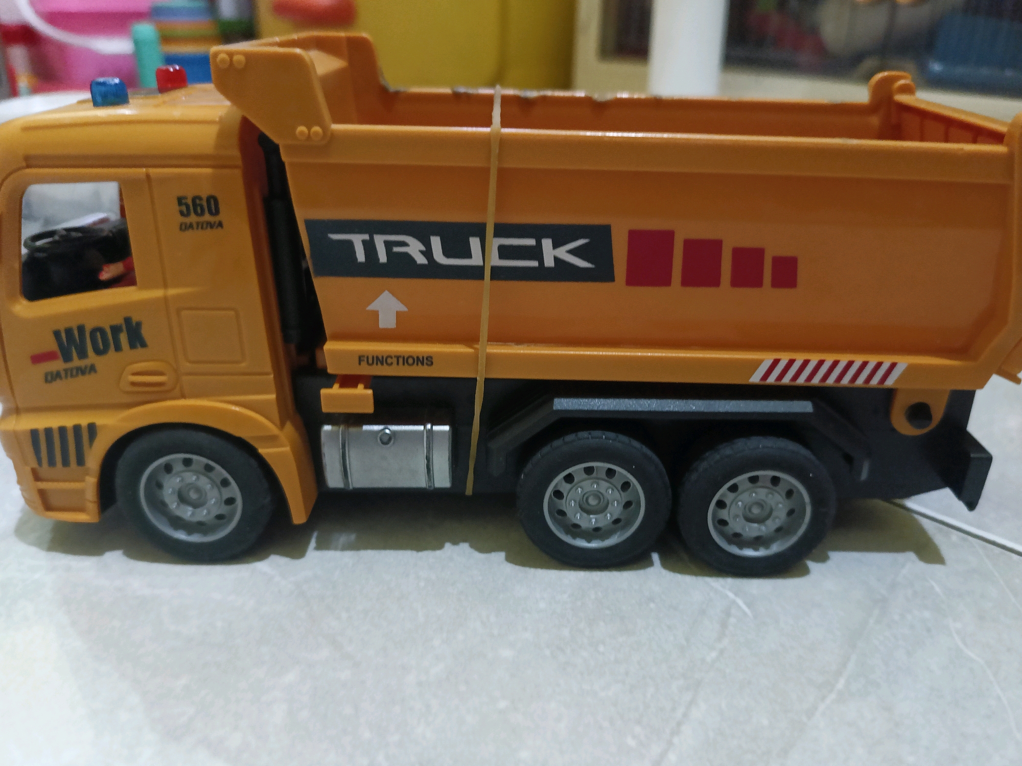 A yellow toy dump truck is shown from a side angle, with its bed raised. The truck has "TRUCK" written on its side in white block letters within a black rectangular background. Below "TRUCK" is a white upward arrow. There are red square graphics to the right of "TRUCK". The truck also has "Work" written on the side of the cab in white lettering with a red dash to the left of it. Above the word "Work" on the cab is a number "560" in black with "DATONA" written below it in smaller black text. The truck has two red lights and one blue light on its roof. The wheels are black with silver rims. A rubber band is stretched vertically across the bed of the truck. The truck is sitting on a light-colored, textured surface, possibly a table or floor. In the background, out of focus, are colorful objects, suggesting a cluttered environment. The image does not provide any information about the city of Tangerang, Indonesia.