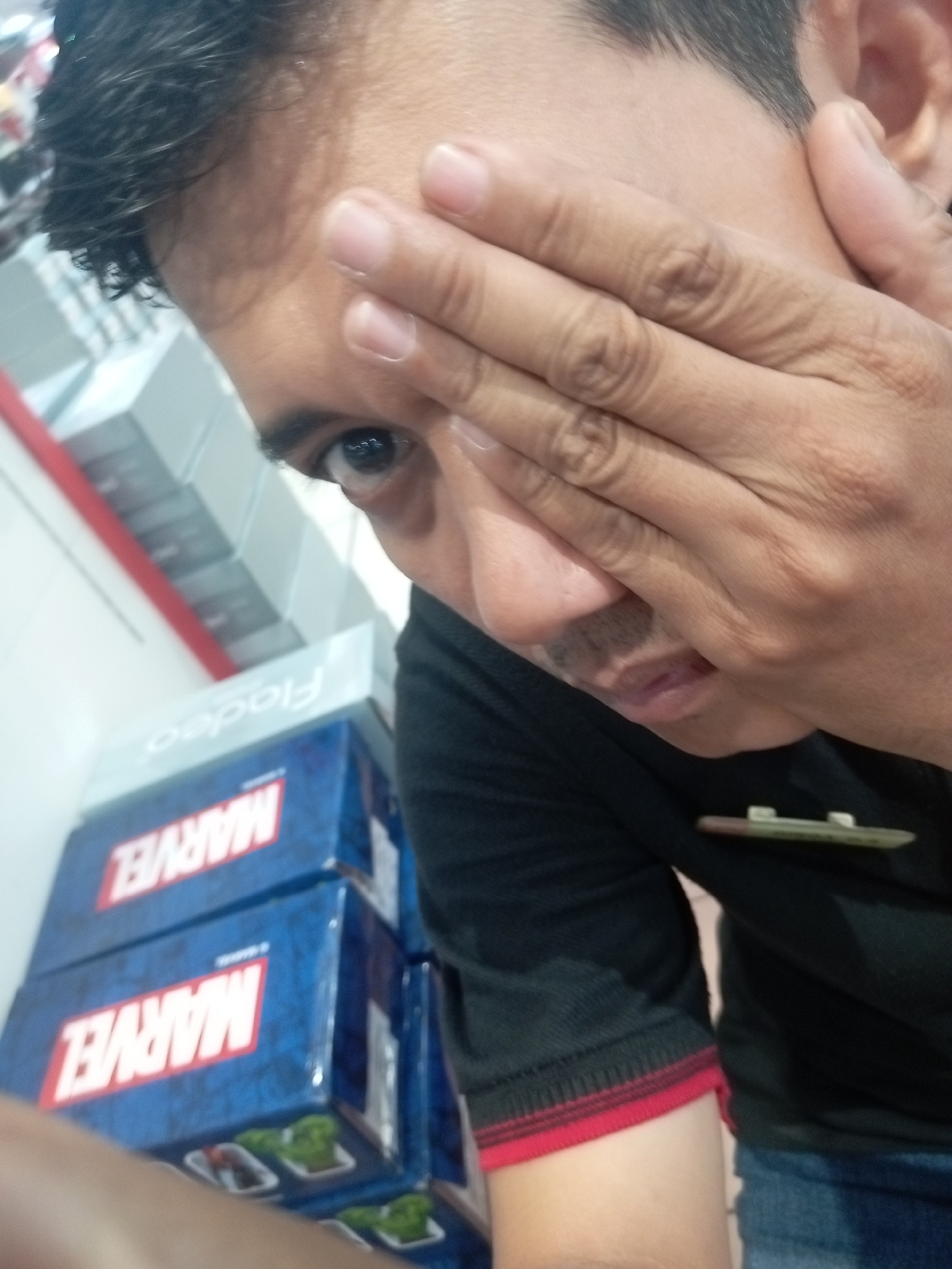 In Batam, Indonesia, a close-up, eye-level shot shows a person covering their face with their right hand, with only one eye visible. The person is wearing a black shirt with red trim on the sleeve and blue jeans. In the background, stacked blue boxes with the "MARVEL" logo are visible, along with a light blue sign that reads "Fiadeo." The setting appears to be an indoor retail environment.