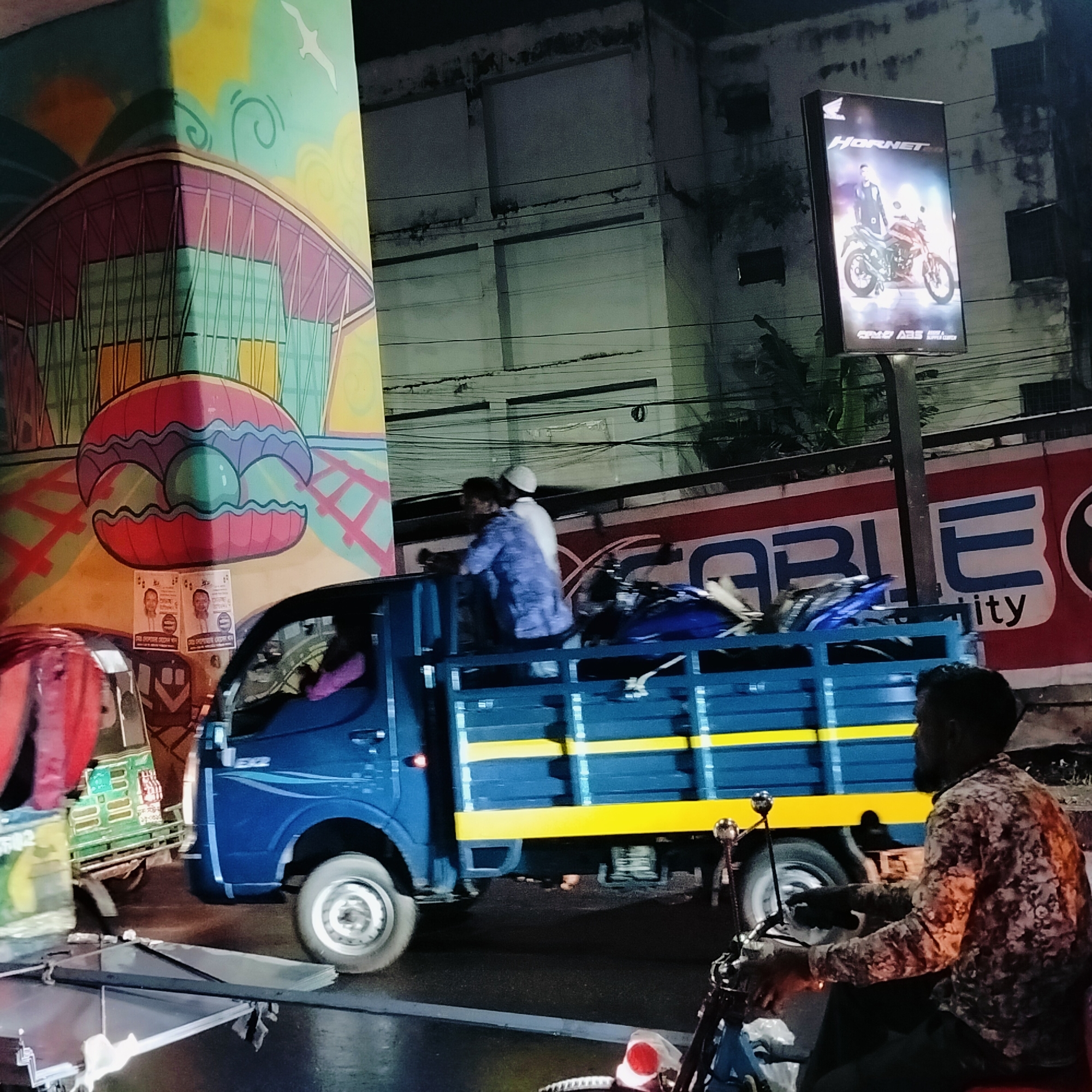The image captures a bustling street scene at night in Dhaka, Bangladesh. A blue Tata Ace truck with yellow accents moves along a street; two men are visible in the open bed of the truck, along with what appears to be a dismantled motorcycle. To the left of the truck is a CNG auto-rickshaw, a common mode of transportation in Dhaka. To the right of the truck, a man is seated on a motorcycle. In the background, a building facade features an advertisement for a Honda Hornet motorcycle. A nearby building wall displays colorful street art, depicting a clamshell and a building skyline. Also, posters are affixed to the street art. The overall scene conveys the chaotic energy of urban traffic in Dhaka.