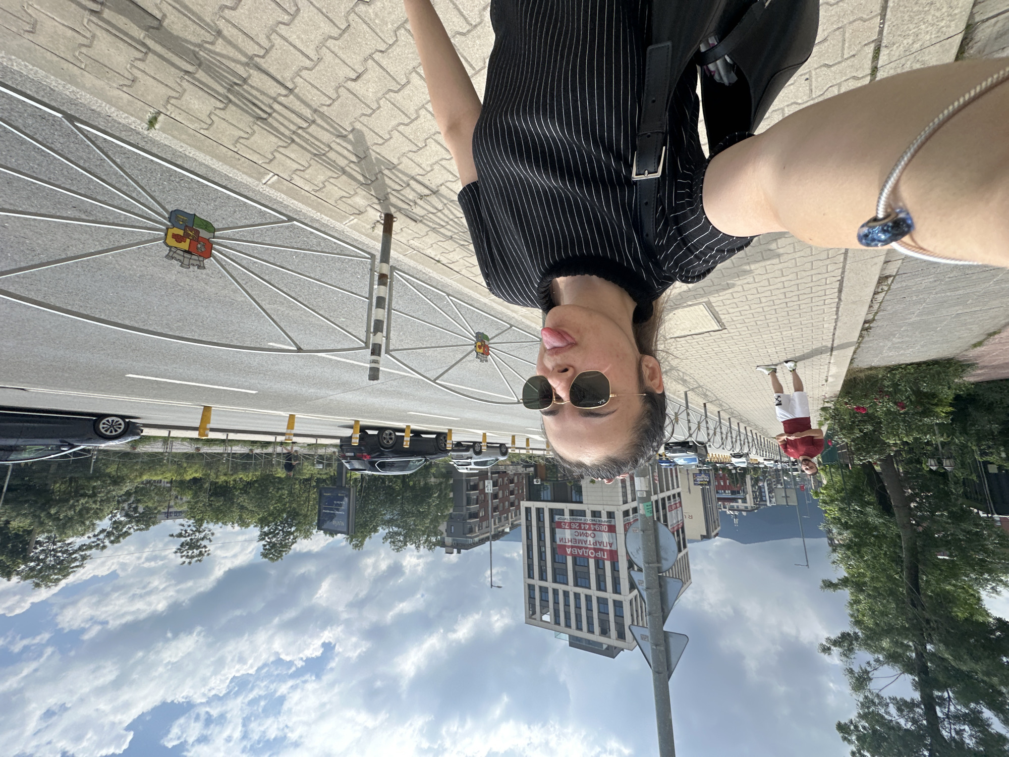 An upside-down selfie photograph captures a young woman on a city sidewalk in Sofia, Bulgaria, during daytime under a partly cloudy sky. The woman, the primary subject, is positioned centrally in the frame. She has dark hair, wears sunglasses, a black and white striped top, and a silver-colored bracelet with a blue charm on her right wrist. Her expression is playful, with her tongue slightly extended. A black shoulder bag is visible on her left side.
Behind her, the scene unfolds with a paved sidewalk bordering a road with traffic markings. Several cars are visible, some parked, others on the move. A prominent feature in the road is a decorative street medallion, possibly resembling a bicycle wheel, with a colorful crest or coat of arms at its center. A black and white striped pole and a street sign are also present.
In the background, a mix of modern and older multi-story buildings line the street. One building features a large red banner with Cyrillic text, reading "ПРОДАВА" (Sells/For Sale), "ОФИС" (Office), "АПАРТАМЕНТИ" (Apartments), and "ДИРЕКТНО ОТ ИНВЕСТИТОР" (Directly from Investor), along with a phone number (0894 44 26 75). Lush green trees and bushes are scattered along the street, and distant mountains are visible under the cloud-dotted sky.
Another person, dressed in a red t-shirt and white shorts, stands on the sidewalk further down the street. The bright lighting suggests it is a clear day, despite the presence of clouds. The overall setting is an active urban environment.