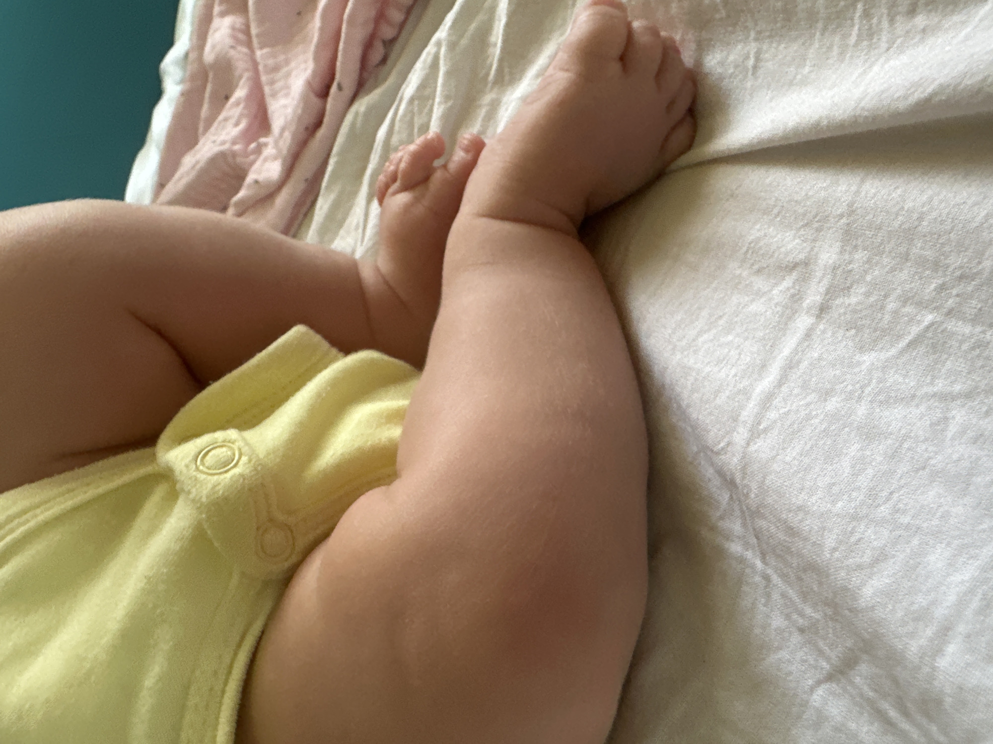 The image is a close-up shot of a baby's legs and feet resting on a white, textured surface, likely a bedsheet. The baby is wearing a light yellow onesie. In the background, a pink blanket with a subtle pattern is visible, and a teal-colored wall can be seen further back. The lighting is soft, highlighting the texture of the baby's skin and the fabric. There is no identifiable information or context related to Plovdiv, Bulgaria, in the image.