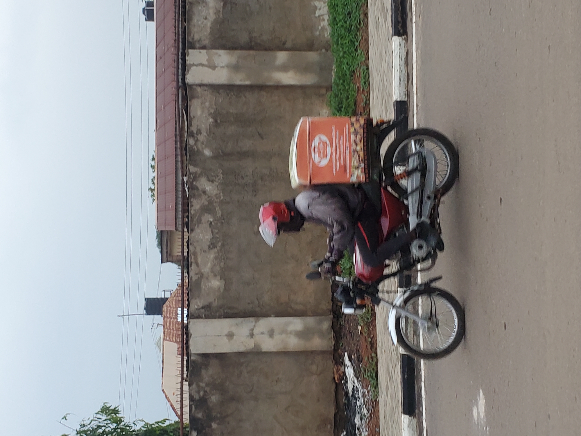 A person is seen riding a red motorcycle on a paved road under an overcast sky. The rider, dressed in a dark jacket and pants, wears a red helmet with a white visor. A large, rectangular orange delivery box is securely mounted on the rear of the motorcycle. The box features a circular logo with unreadable text or graphics.
The motorcycle is positioned near the edge of the road, which has a white line and a black and white painted curb. Beyond the curb, there's a strip of brown dirt with some green grass. A tall, textured concrete wall dominates the background, and behind it, parts of buildings with reddish-brown corrugated metal roofs are visible. Power lines traverse the light grey sky above the buildings. The scene depicts a typical street in an urban or semi-urban area, consistent with the provided location context of Narayi, Nigeria. The time of day appears to be daytime, possibly during cloudy weather.