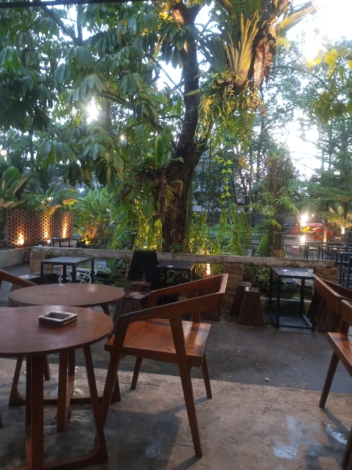 This image depicts an empty outdoor cafe or restaurant patio in Pontianak, Indonesia, during the evening. The foreground features a concrete floor with several vacant wooden tables and chairs. A round wooden table in the immediate foreground displays a metal ashtray, while another round wooden table behind it holds a dark-colored smartphone. Adjacent to these are modern wooden chairs with distinct angled designs.
The middle ground presents additional seating options, including square black metal tables and more wooden chairs, interspersed with dark wooden stumps used as stools or decor. A low, light-colored brick wall with integrated uplighting separates this area from a dense garden space. A dark, possibly chalkboard-style, sign stands near one of the square tables.
The background is characterized by abundant, lush green foliage dominated by large trees, ferns, and various other plants. String lights are visible within the canopy, and additional ground-level accent lighting illuminates the vegetation and brick wall. A bright light source, possibly the setting sun or a powerful artificial light, pierces through the leaves in the upper left quadrant. Metal fencing or grating is discernible through the dense greenery further back, indicating an enclosed or semi-enclosed outdoor setting. No individuals are present.