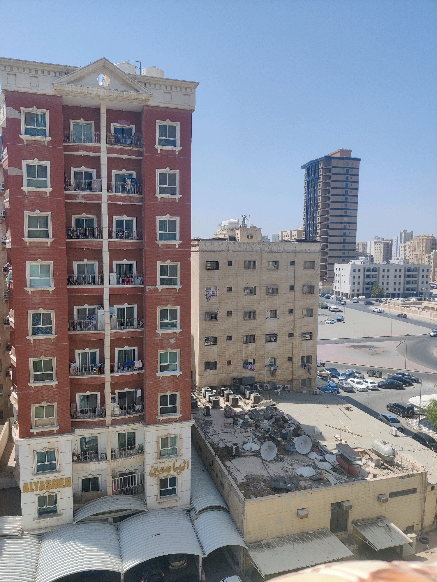 The image shows an aerial view of Salmiya, Kuwait. In the foreground, a multi-story residential building with red brick facade and white trim stands prominently. Balconies with railings are visible on multiple floors, with some showing clothes drying. The building has a central white section with Arabic lettering reading "AL YASMEN". Adjacent to this building is a lower, light-colored building with a flat roof covered in satellite dishes and debris. In the background, several other urban buildings of varying heights and architectural styles are visible. A tall, slender skyscraper with blue accents is a notable landmark in the distance. The street below is visible, with several cars parked and driving on a curved road. The sky is clear and blue, indicating a sunny day.