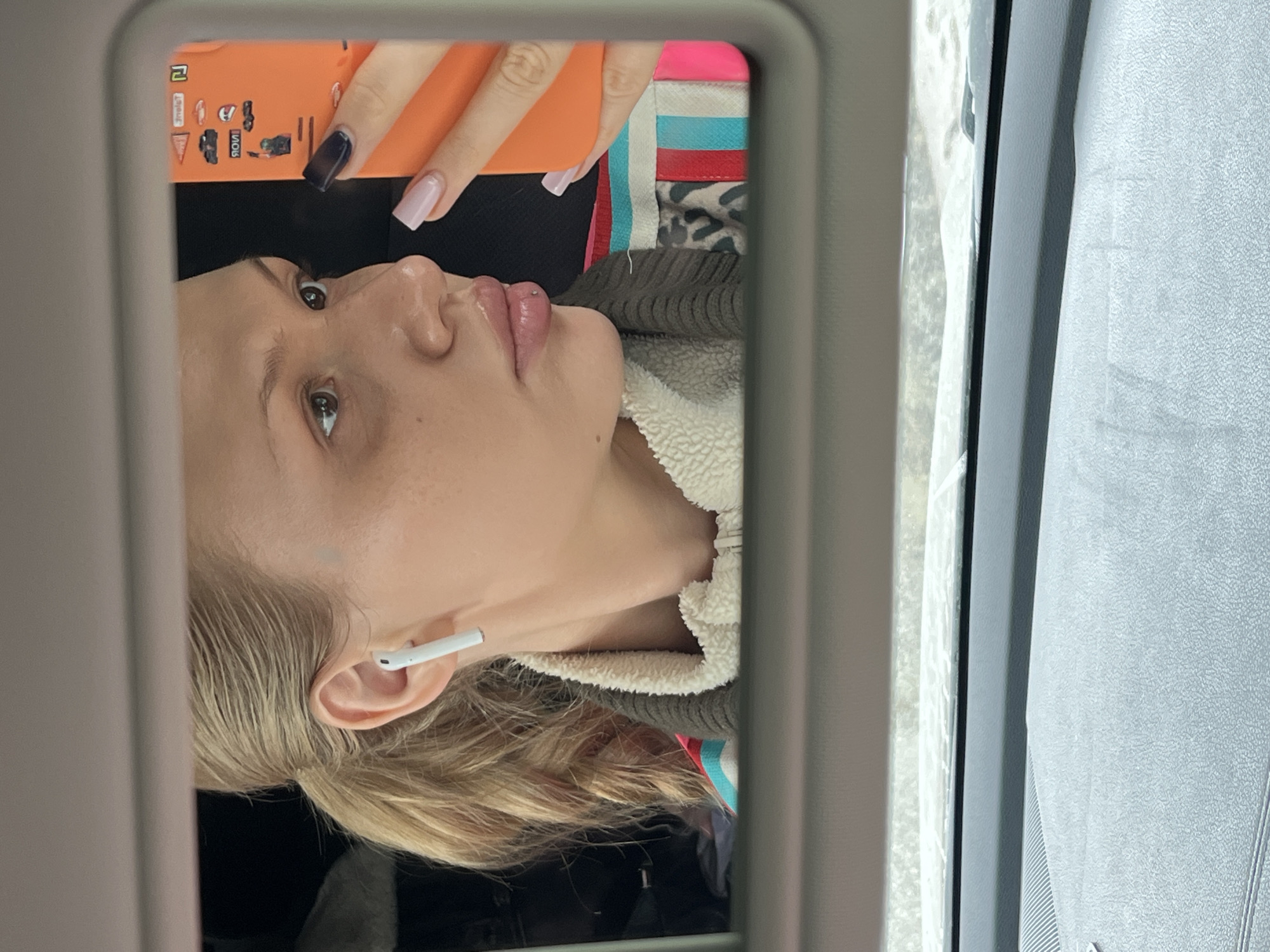 A woman with blonde hair is pictured in a car's vanity mirror. She has an earbud in her left ear and is holding an orange phone. Her nails are manicured, with one painted black and the other a light pink. She has a lip piercing. The car window and visor are visible. The location and city are indeterminable from the image.