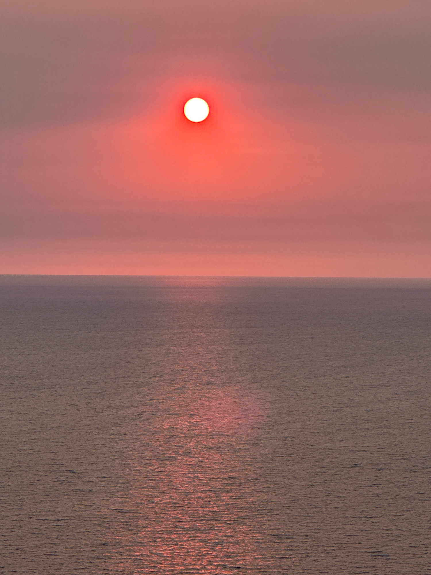 The image shows a seascape at sunset. The sun, a bright red-orange orb, is partially obscured by a hazy pink sky, casting a shimmering reflection on the calm, dark water. The horizon line is sharp and clearly divides the sky from the sea. There are no people or man-made objects visible in the image. The scene is serene and atmospheric, conveying a sense of tranquility. The location in the city of Zagora, Montenegro is not discernible from the visual information provided in the image itself.