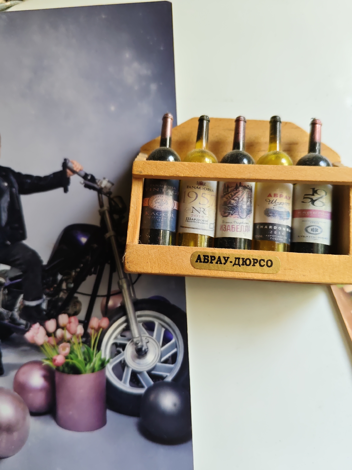 The image shows a close-up of a miniature wine rack containing four small bottles of wine. The rack is made of light-colored wood and is positioned in front of a backdrop that features a blurred image of a person riding a motorcycle, with pink flowers and metallic spheres in the foreground. The wine bottles have labels with Cyrillic text and what appear to be vintage designs. One label prominently displays "АВРАУ-ДЮРСО". The scene is indoors, likely a home or a display, with neutral lighting. The overall impression is that of a decorative item or souvenir. The text on the label "АВРАУ-ДЮРСО" refers to Abrau-Durso, a renowned winery located in the Krasnodar Krai region of Russia, not in Rostov-on-Don.